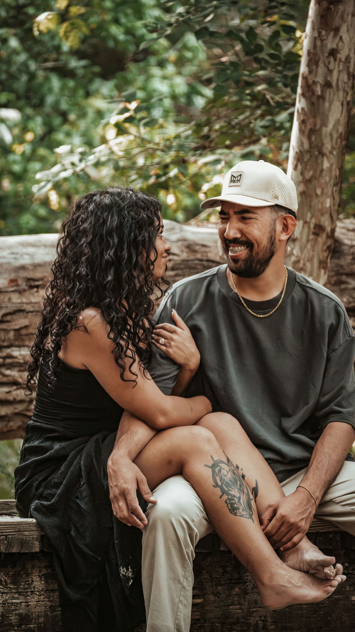 Laughing couple sitting on a rustic log bench in a wooded area, sharing a joyful intimate moment together.