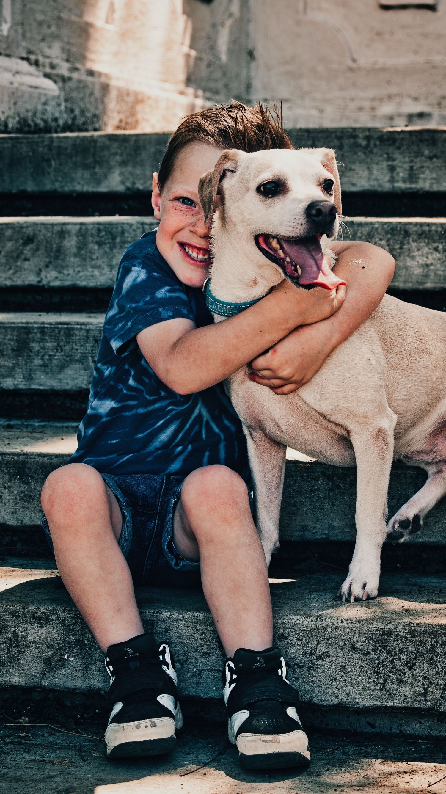 Happy red-haired boy hugging a white dog while sitting on stone steps outdoors. Family Photographer in Pflugerville, Tx