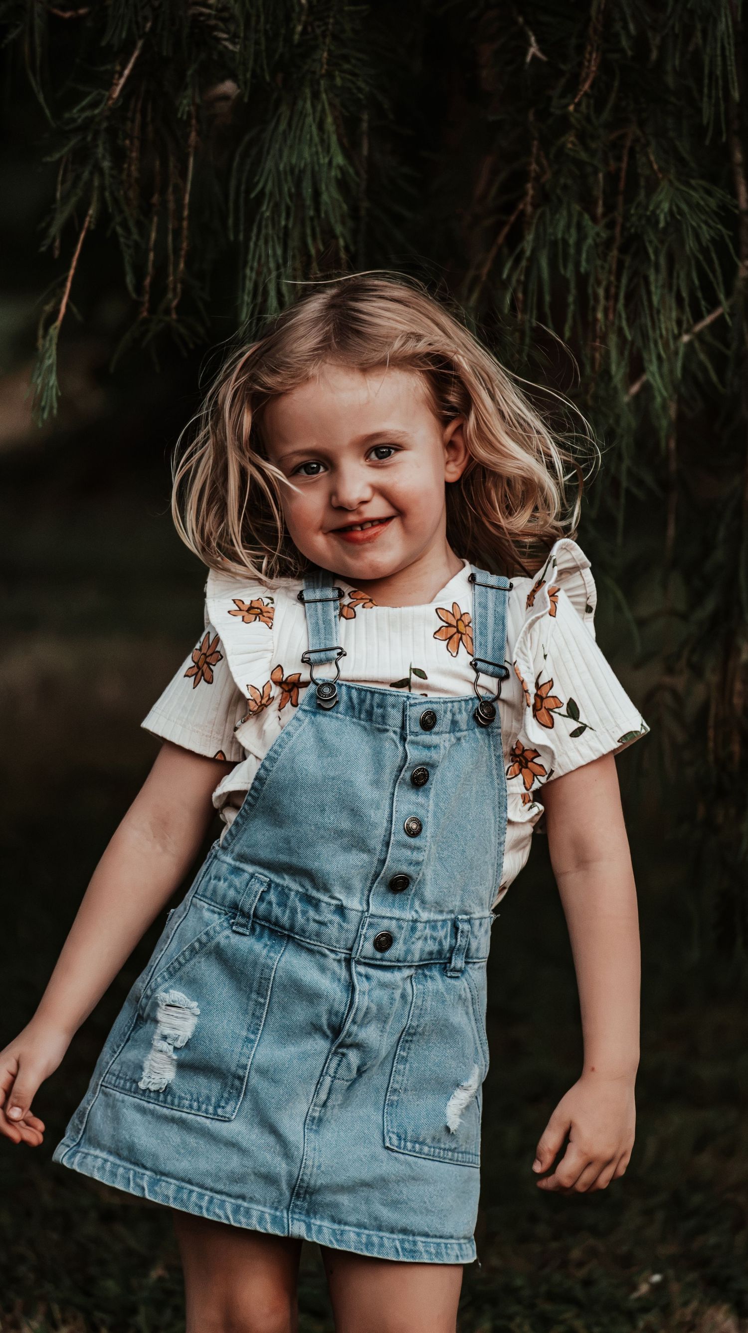 Smiling young girl in denim overalls and floral shirt poses outdoors near green pine trees.