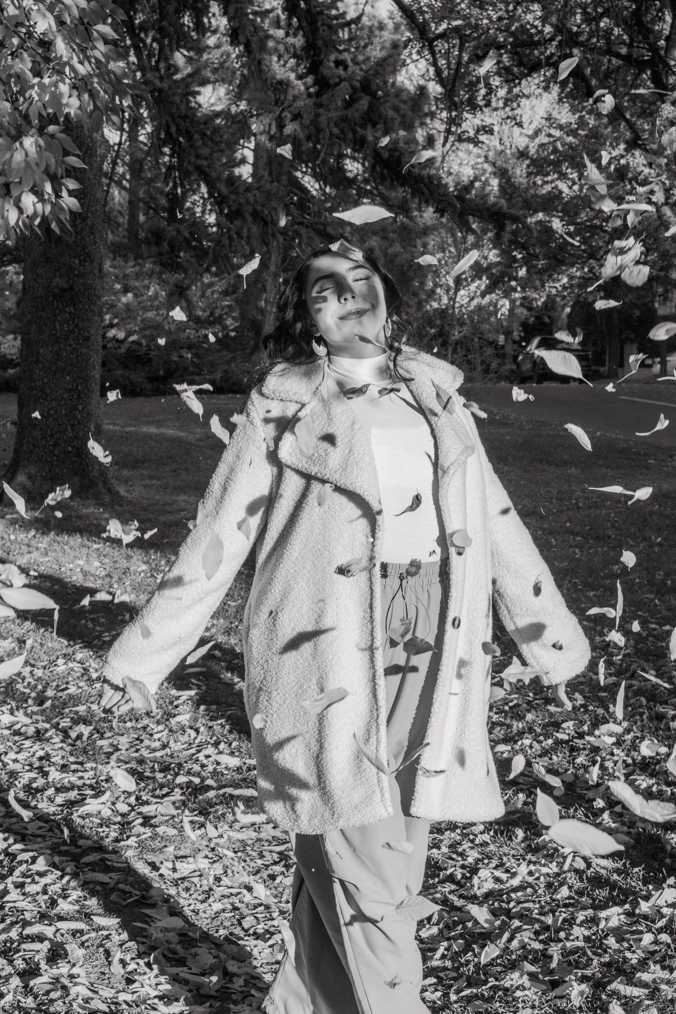 Black and white artistic shot of woman in oversized coat with leaves falling around her for senior photos.