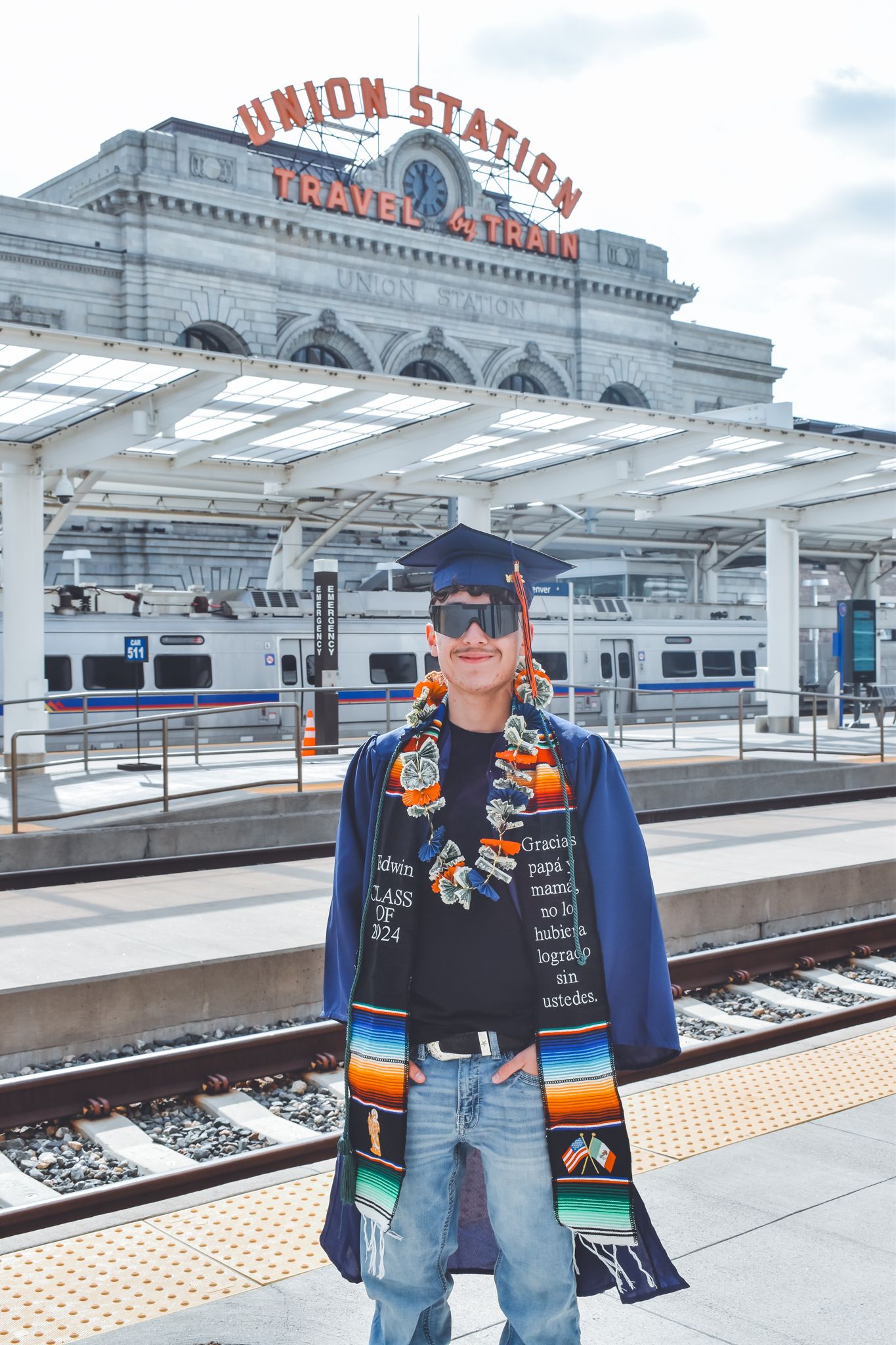 Graduate in blue cap and gown stands on train tracks in front of Denver Union Station for senior photos.
