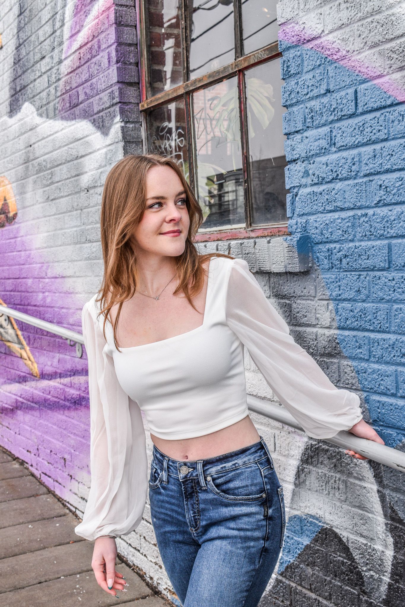 Young woman in white crop top and jeans poses against colorful graffiti wall in urban setting for senior photos.