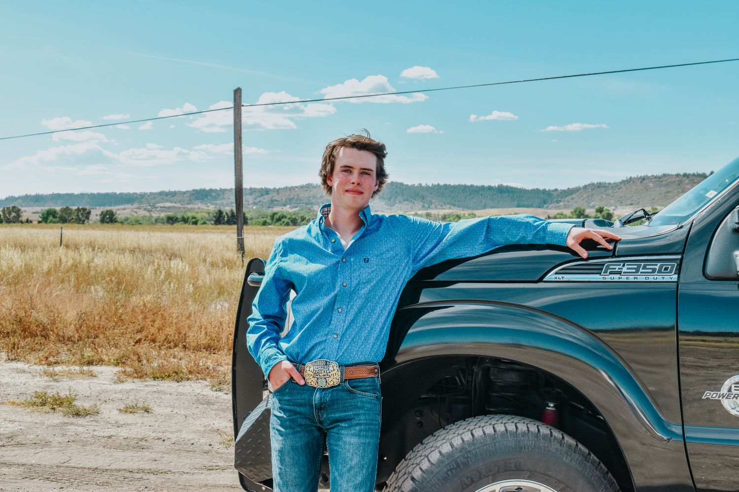 Young man in blue western shirt with belt buckle leaning against a dark green pickup truck outdoors for senior photos.
