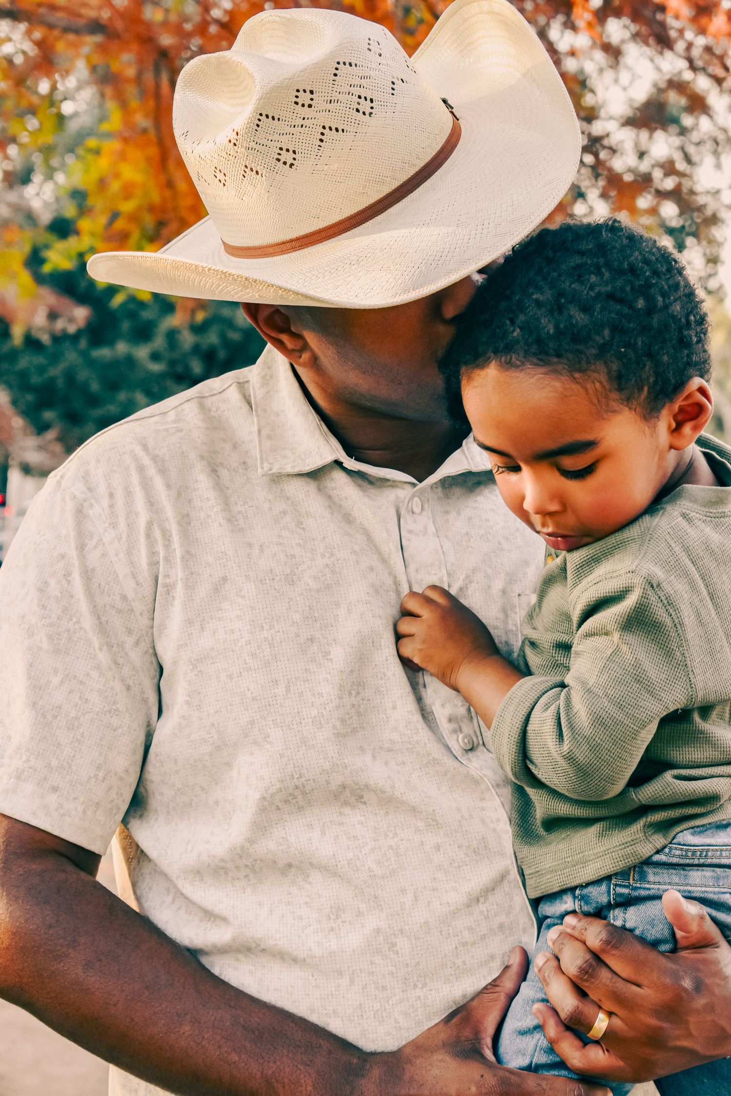 A father wearing a cowboy hat tenderly holds his young son in a warm autumn park scene.