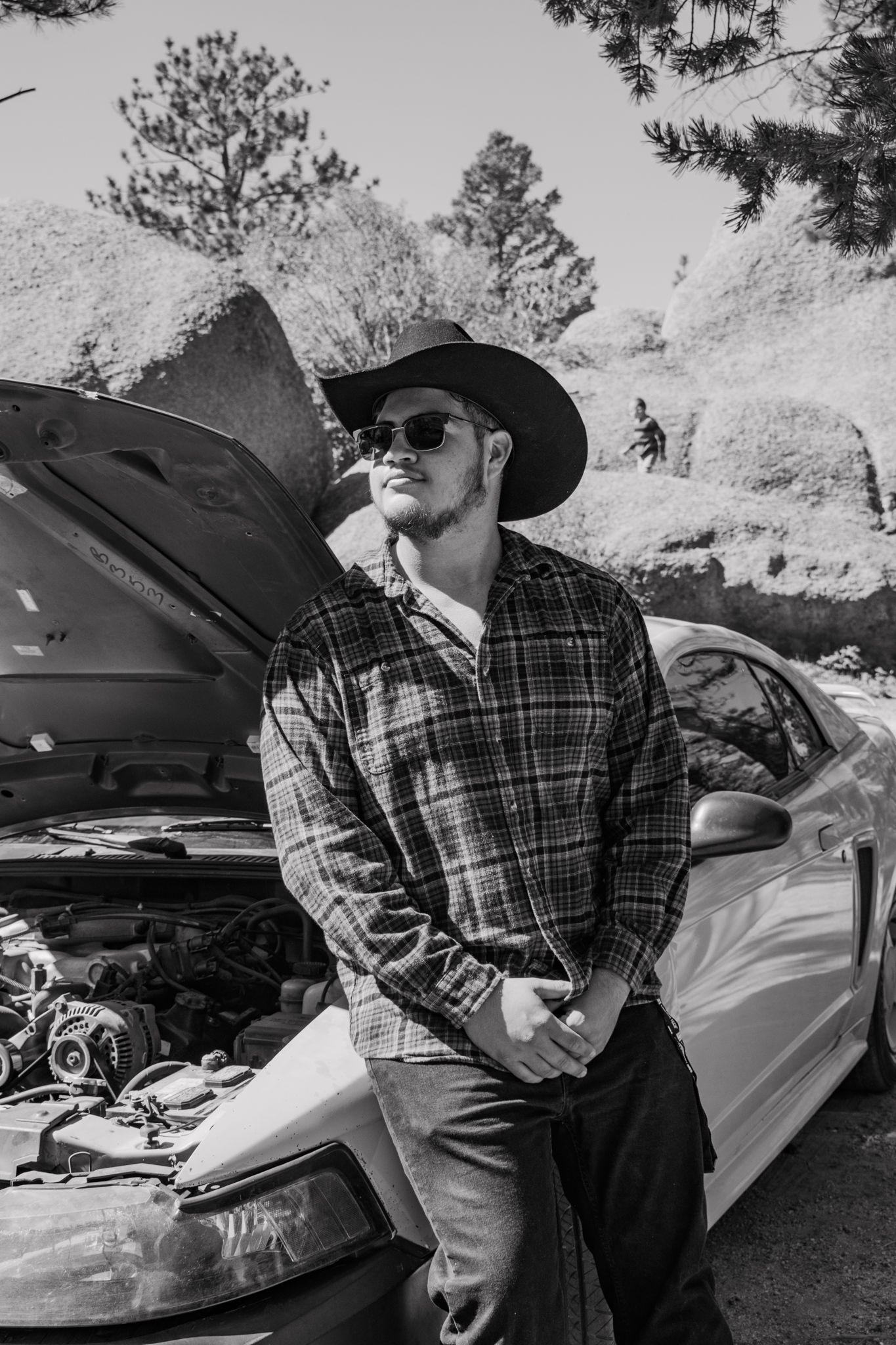 Black and white photo of man in cowboy hat and plaid shirt leaning on car with open hood for senior photos.