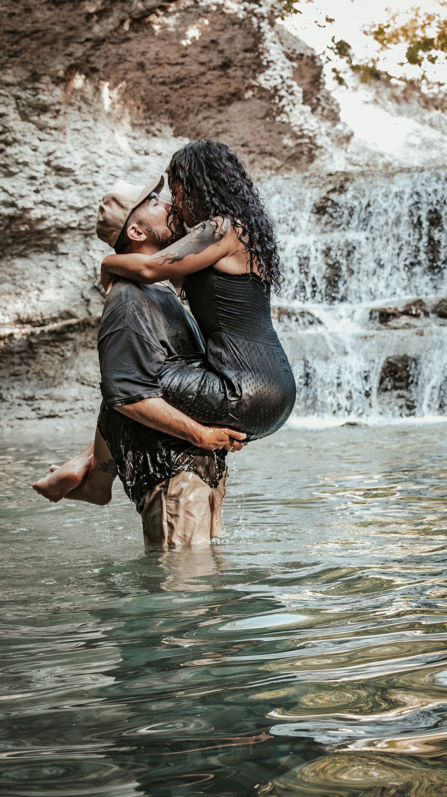 A romantic couple embraces in a waterfall pool, the man lifting the woman in a black dress for a couples session.
