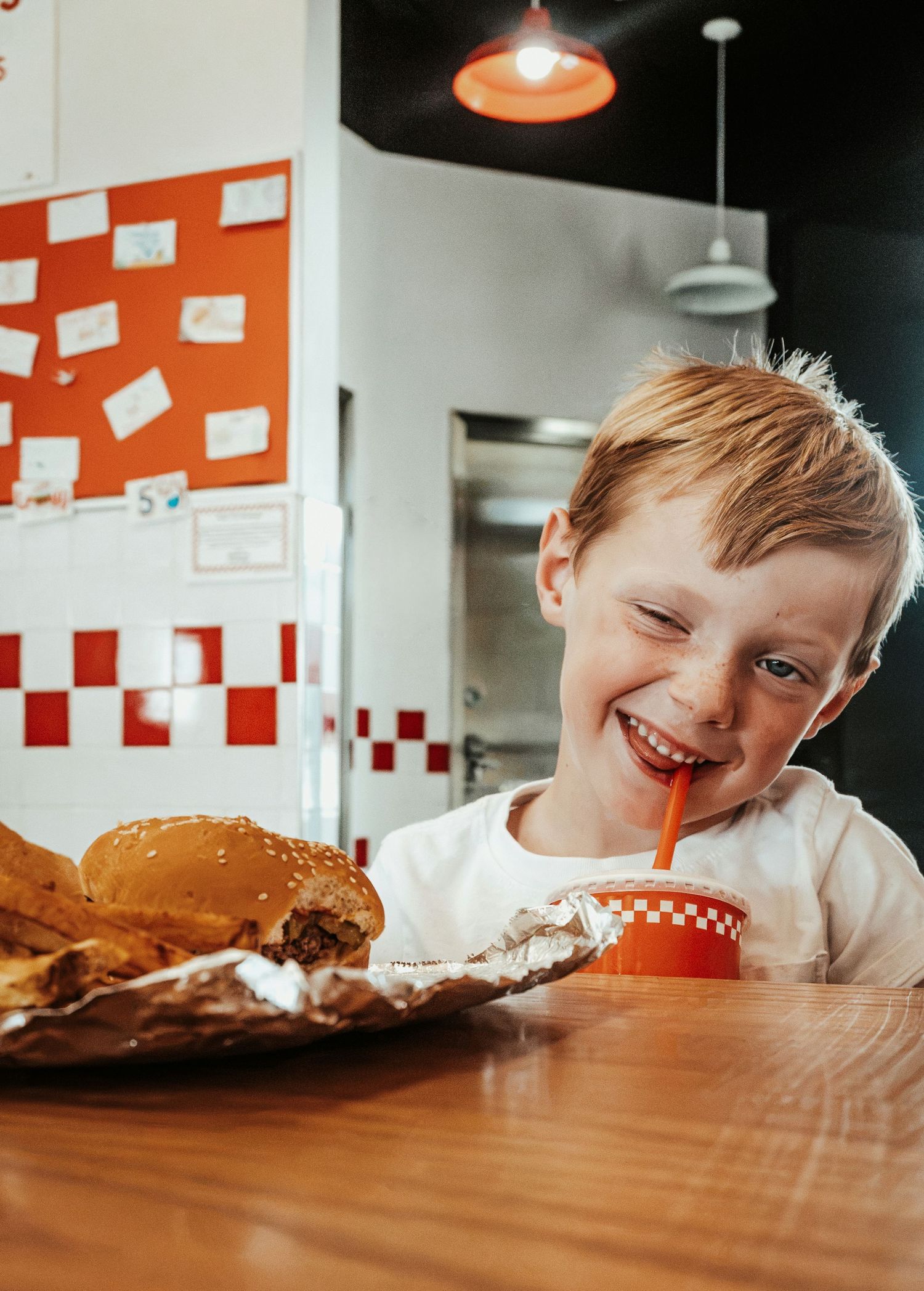 Smiling red-haired boy enjoying a burger and drink at Five Guys restaurant. Family Photographer in Pflugerville, Tx