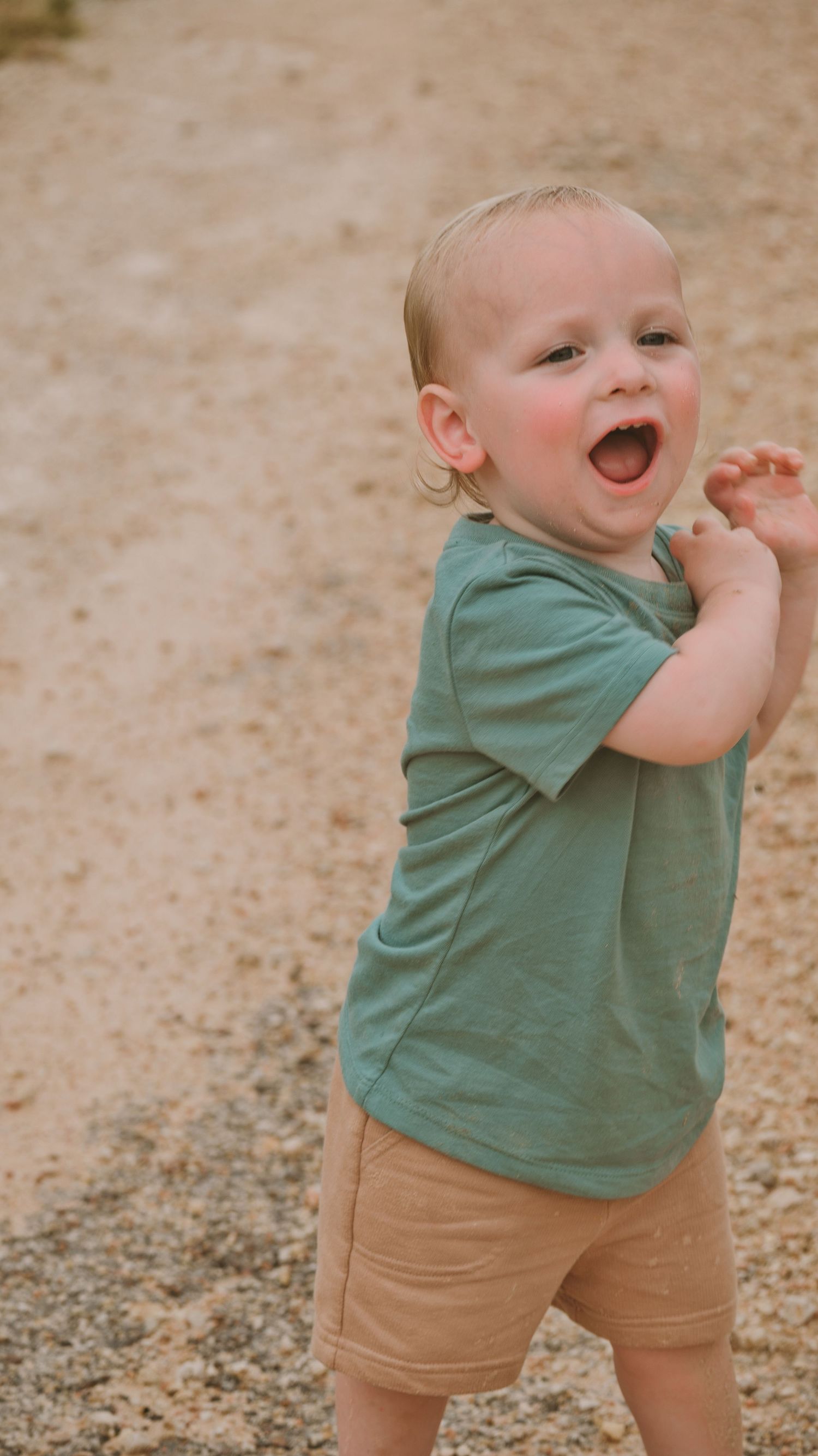 A happy toddler in a green t-shirt and tan shorts laughs and plays on a sandy outdoor path for a family session.