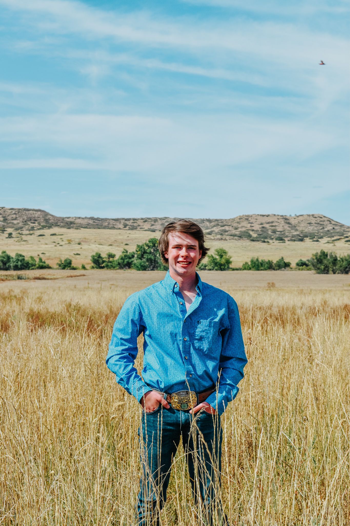 Smiling young man in blue western shirt and decorative belt buckle standing in golden prairie grass field for senior photos.
