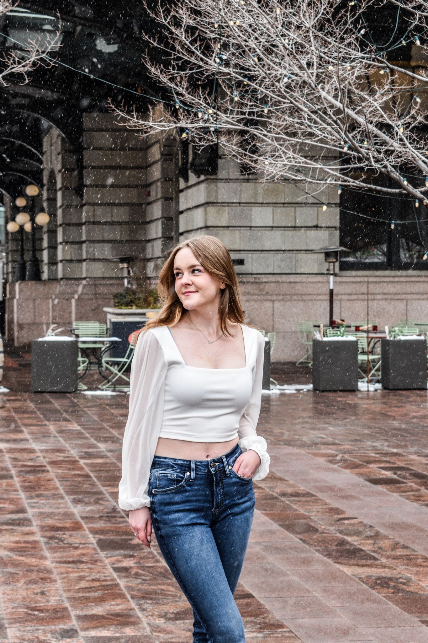 Young woman in white square-neck crop top and jeans posing outdoors in snowy city plaza in winter for senior photos.