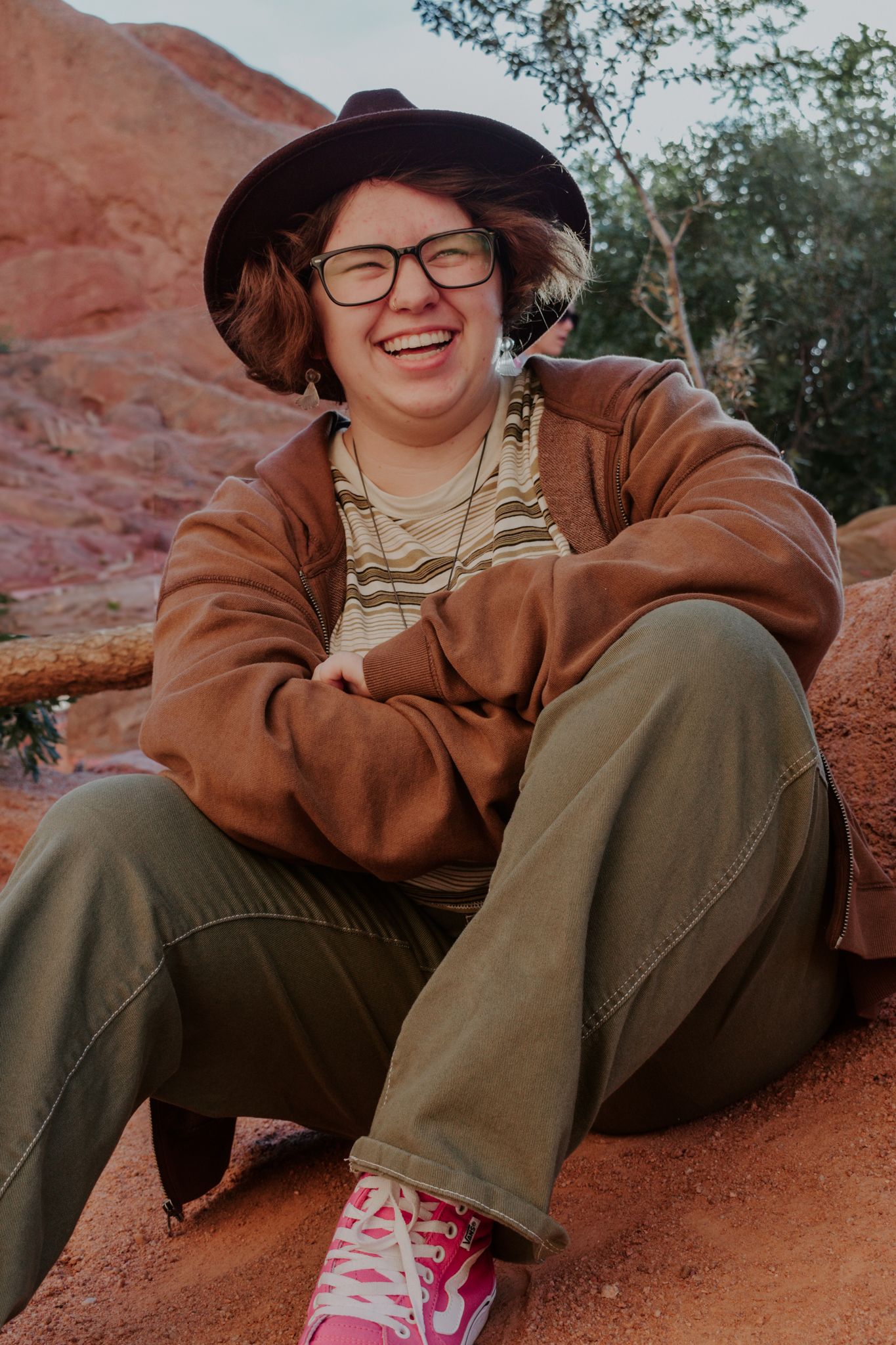 Person sitting on red rocks laughing, wearing a brown jacket, hat, glasses, and pink sneakers for senior photos.