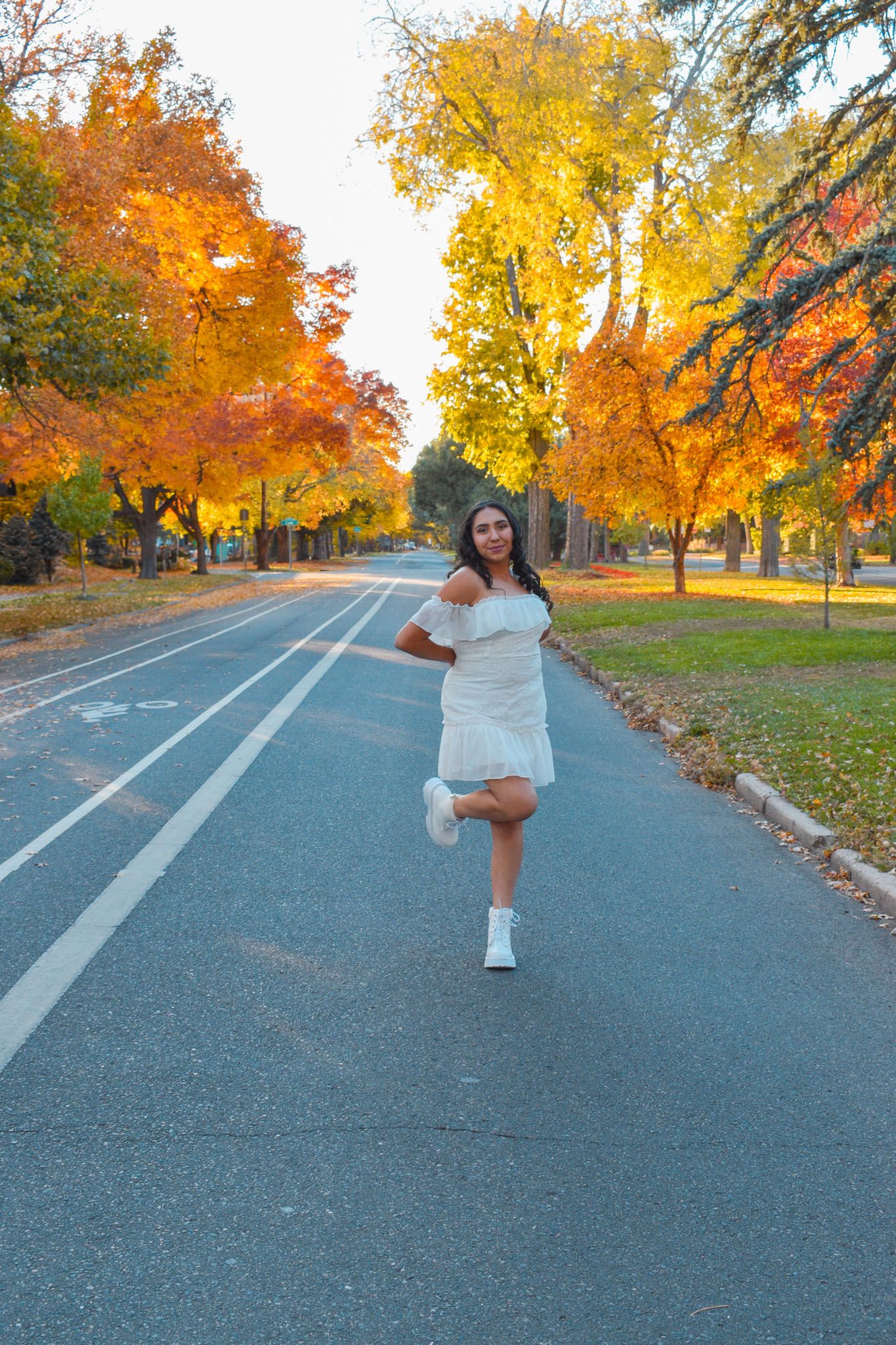 Young woman in white dress playfully posing on a road lined with vibrant fall foliage for senior photos.