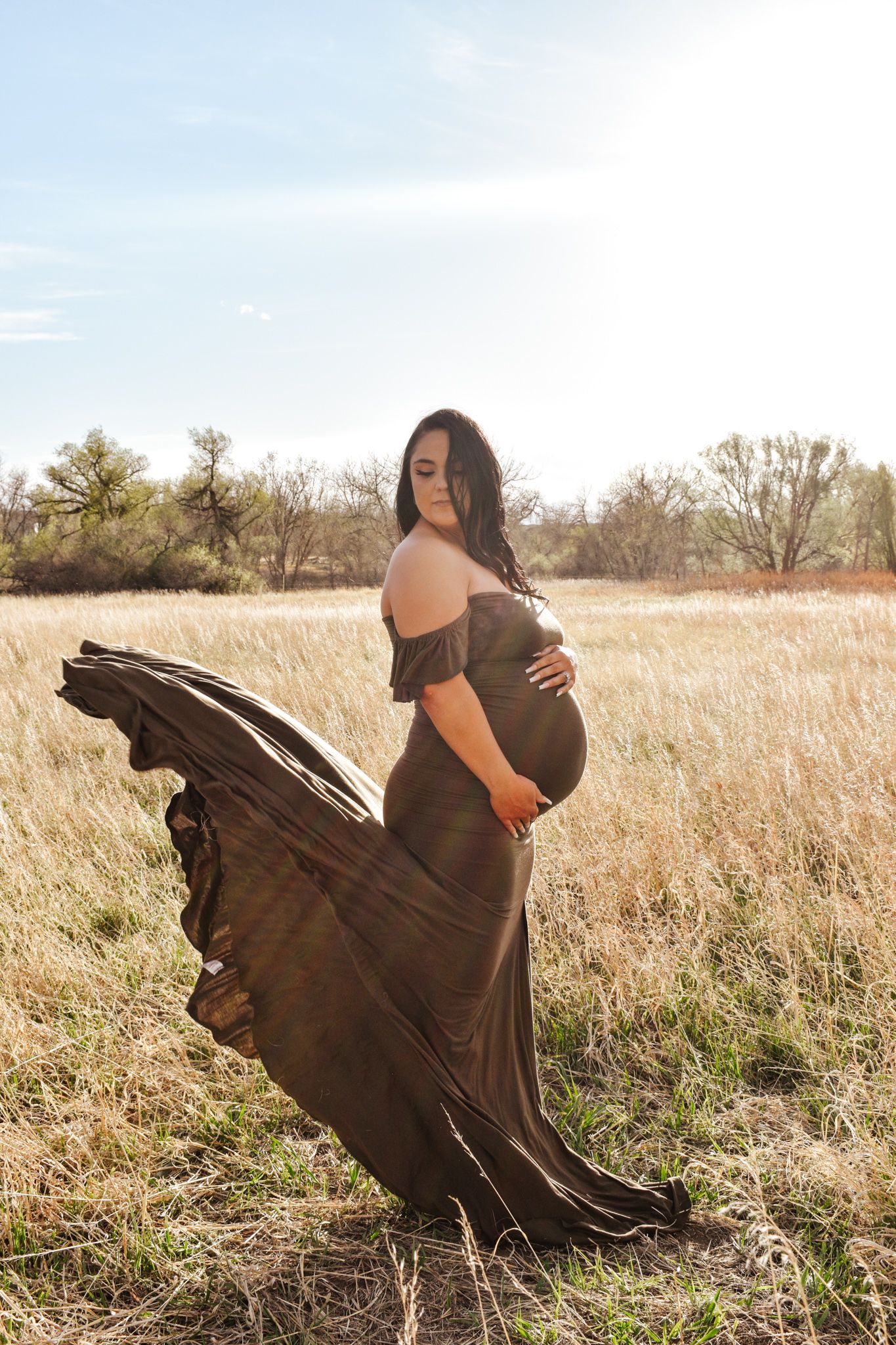 Pregnant woman in flowing olive off-shoulder gown with dress billowing in wind in sunny field.