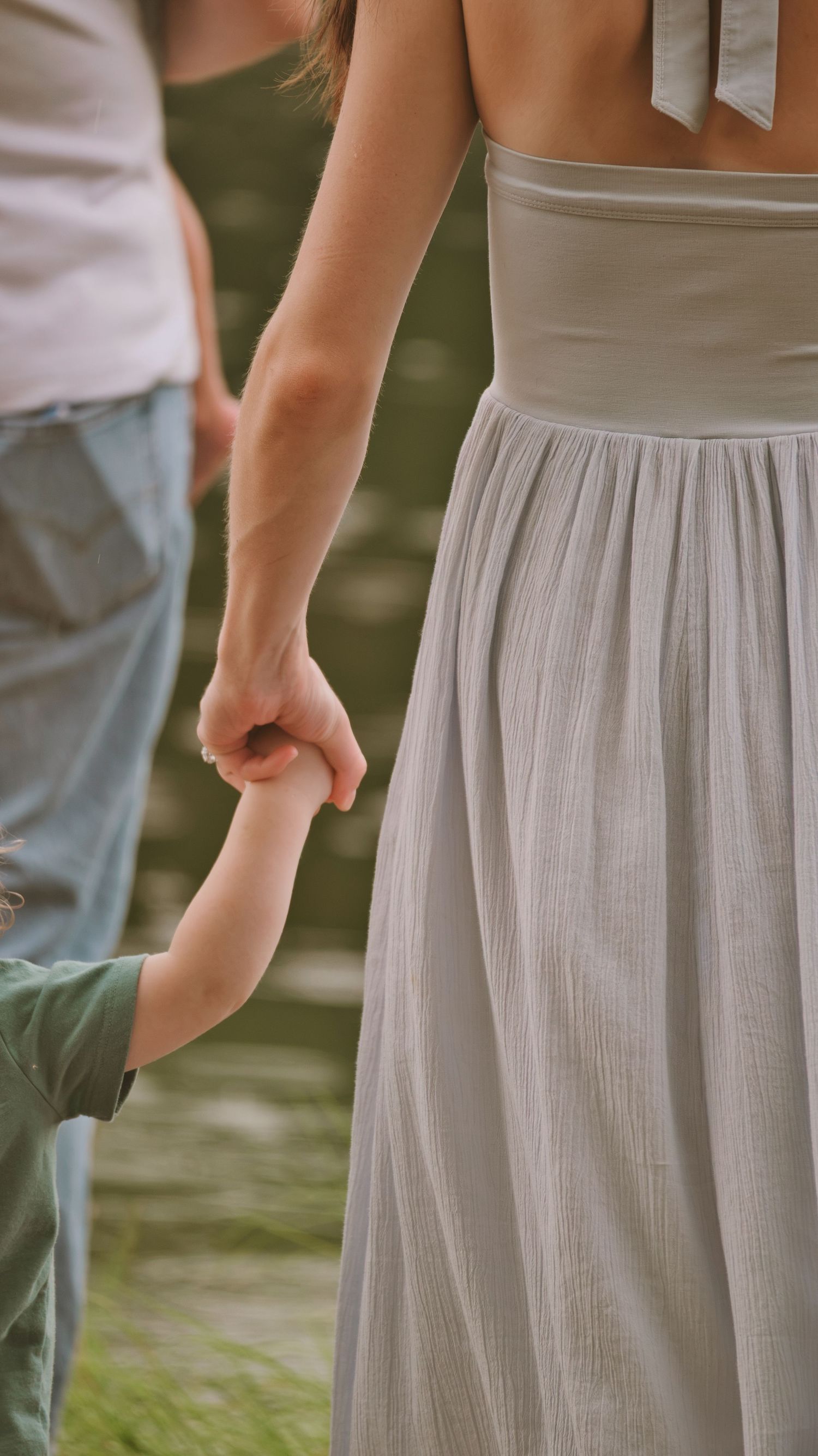 A woman in a gray dress holds a young child's hand while walking outdoors, with a man in jeans nearby.