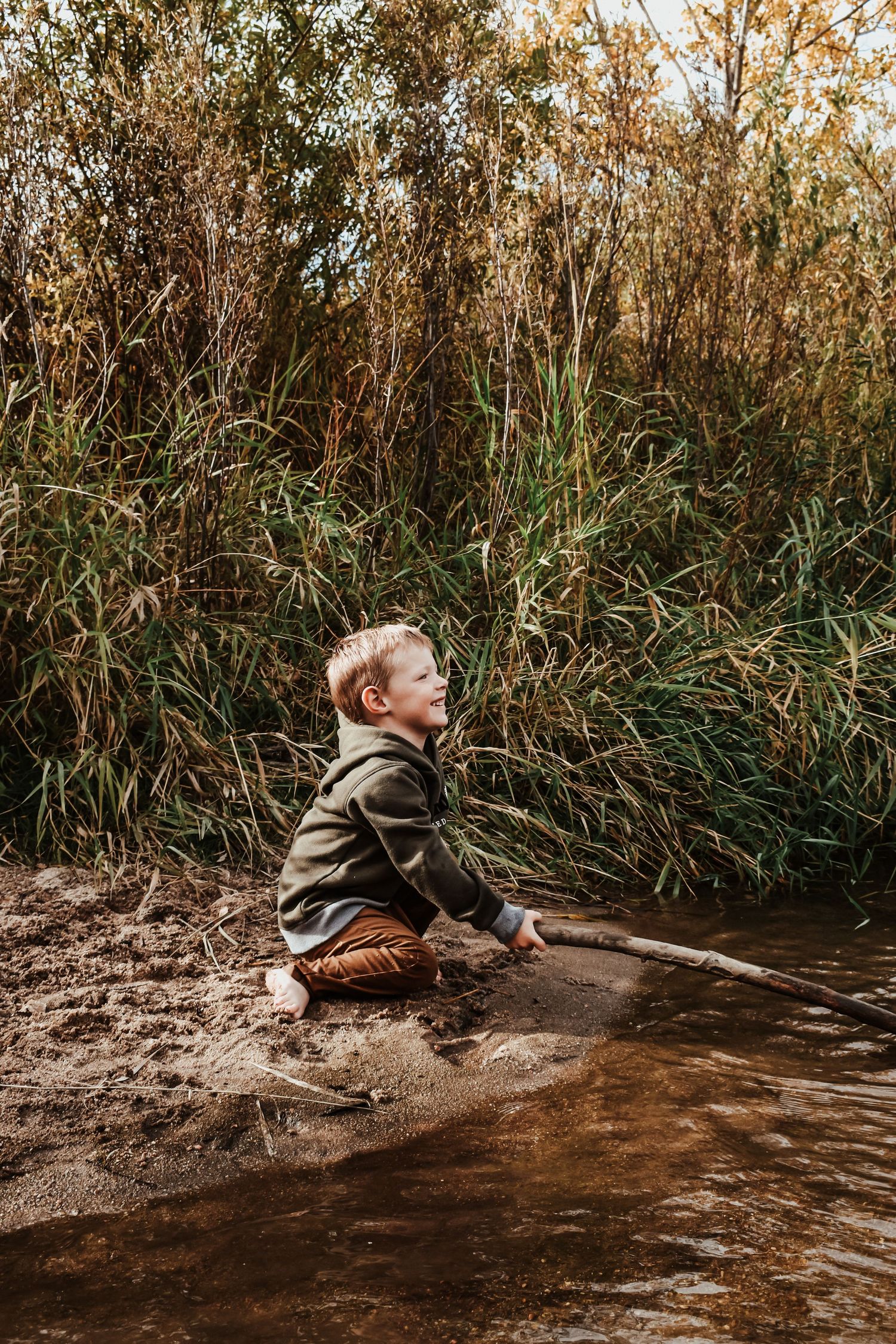 Young boy sitting on a sandy creek bank holding a stick and looking up surrounded by tall reeds.