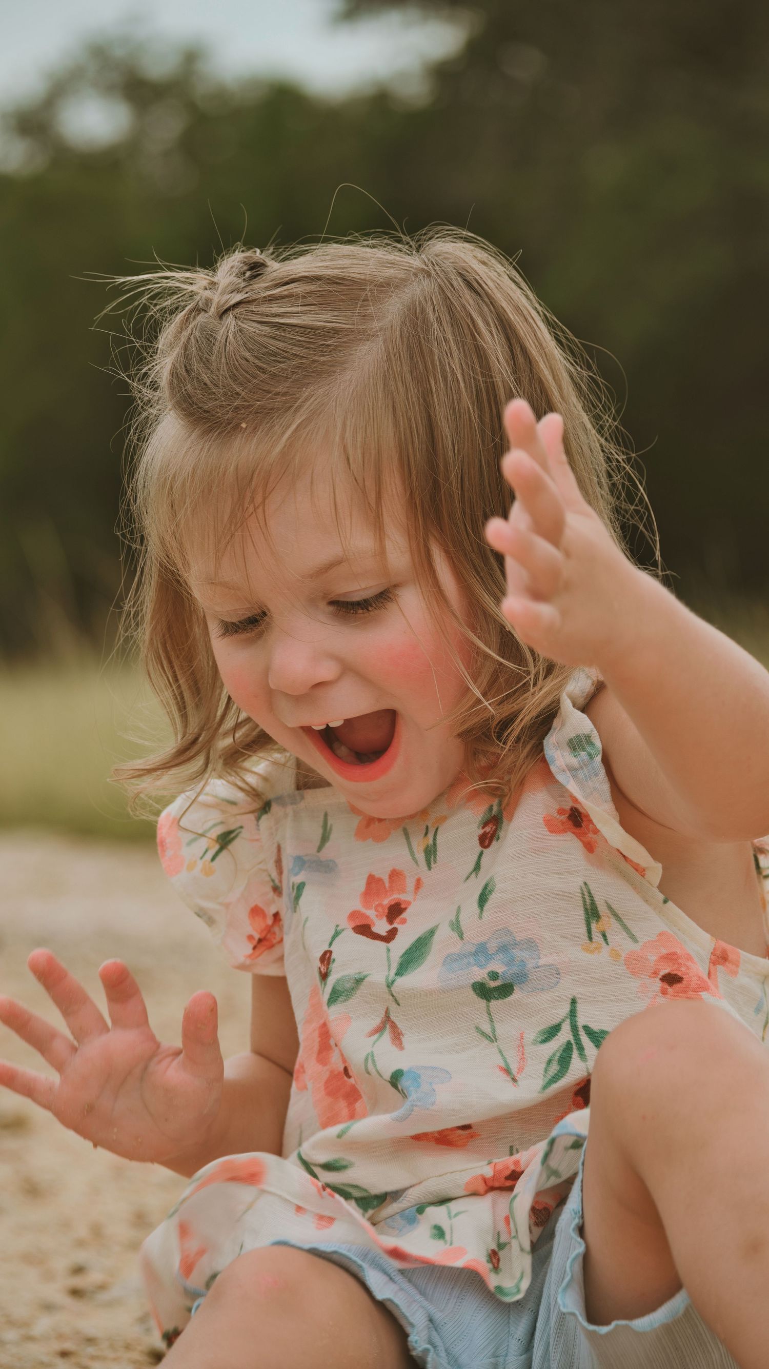 Excited toddler girl in floral dress laughing with hands raised outdoors for a family session.