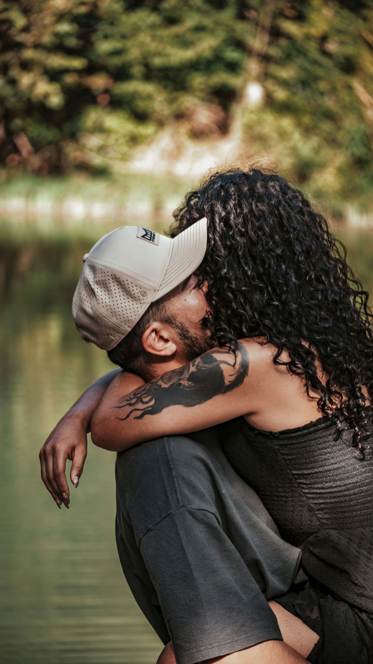 A woman with curly hair embraces a tattooed man wearing a cap by a peaceful riverside.