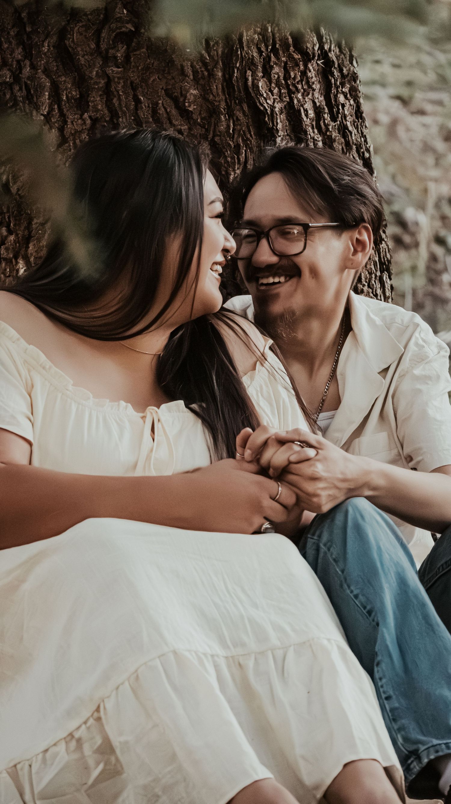 Smiling couple holding hands while sitting against a tree trunk outdoors.