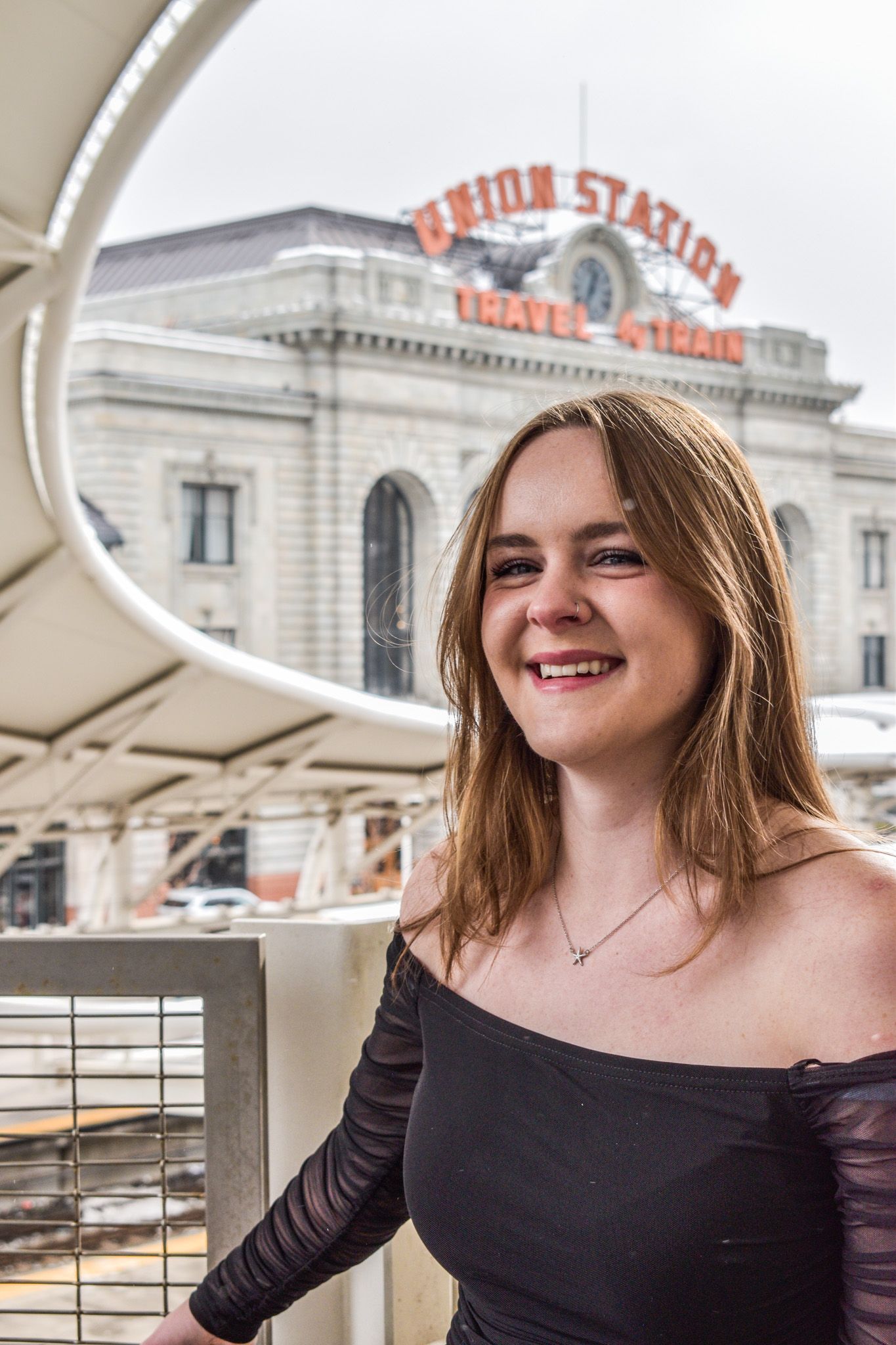Smiling young woman in black off-shoulder dress posing at Denver Union Station Travel Center outdoors for senior photos.