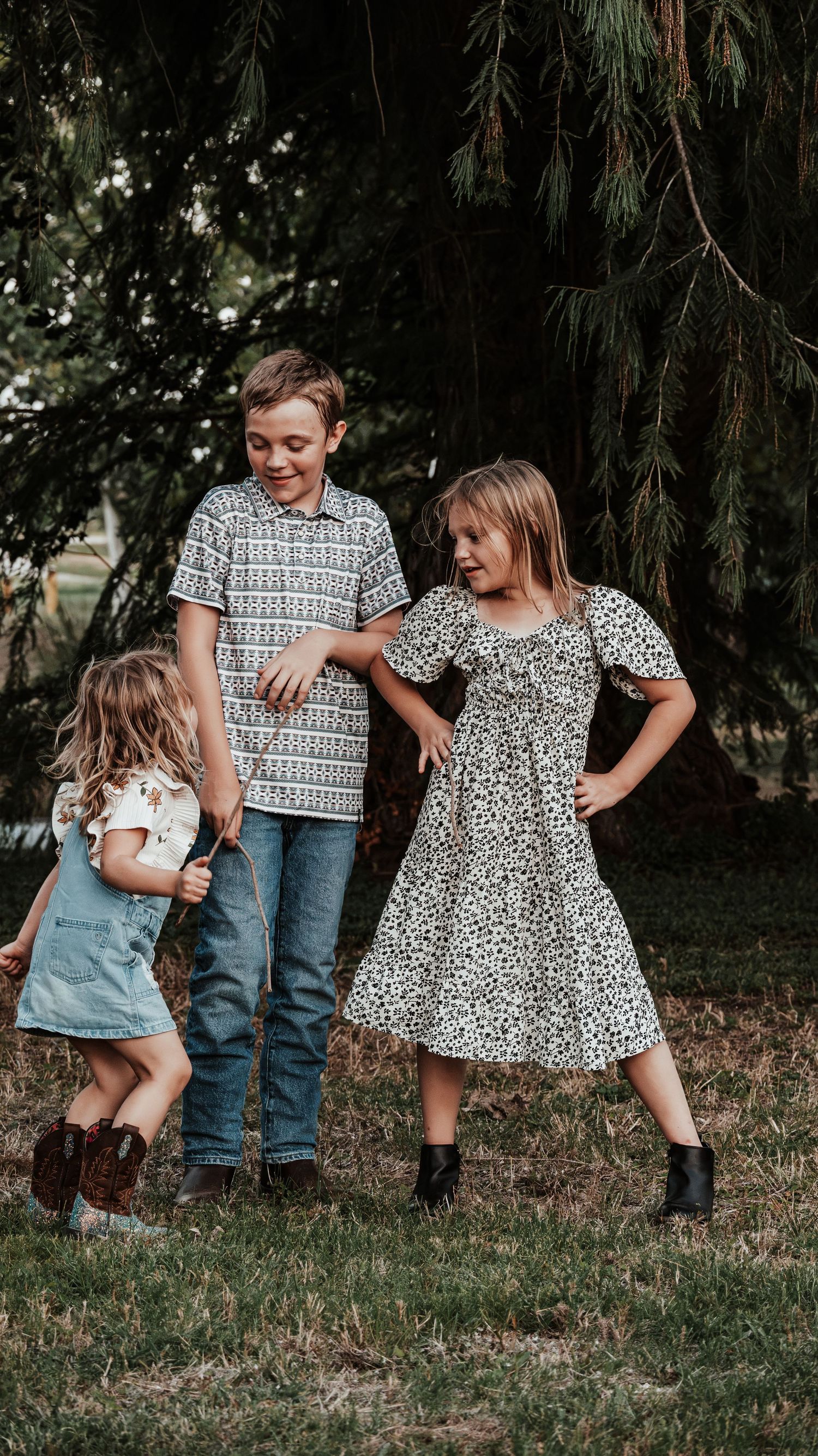 Three siblings playing outdoors under pine trees, older boy and girl watching over toddler sister.