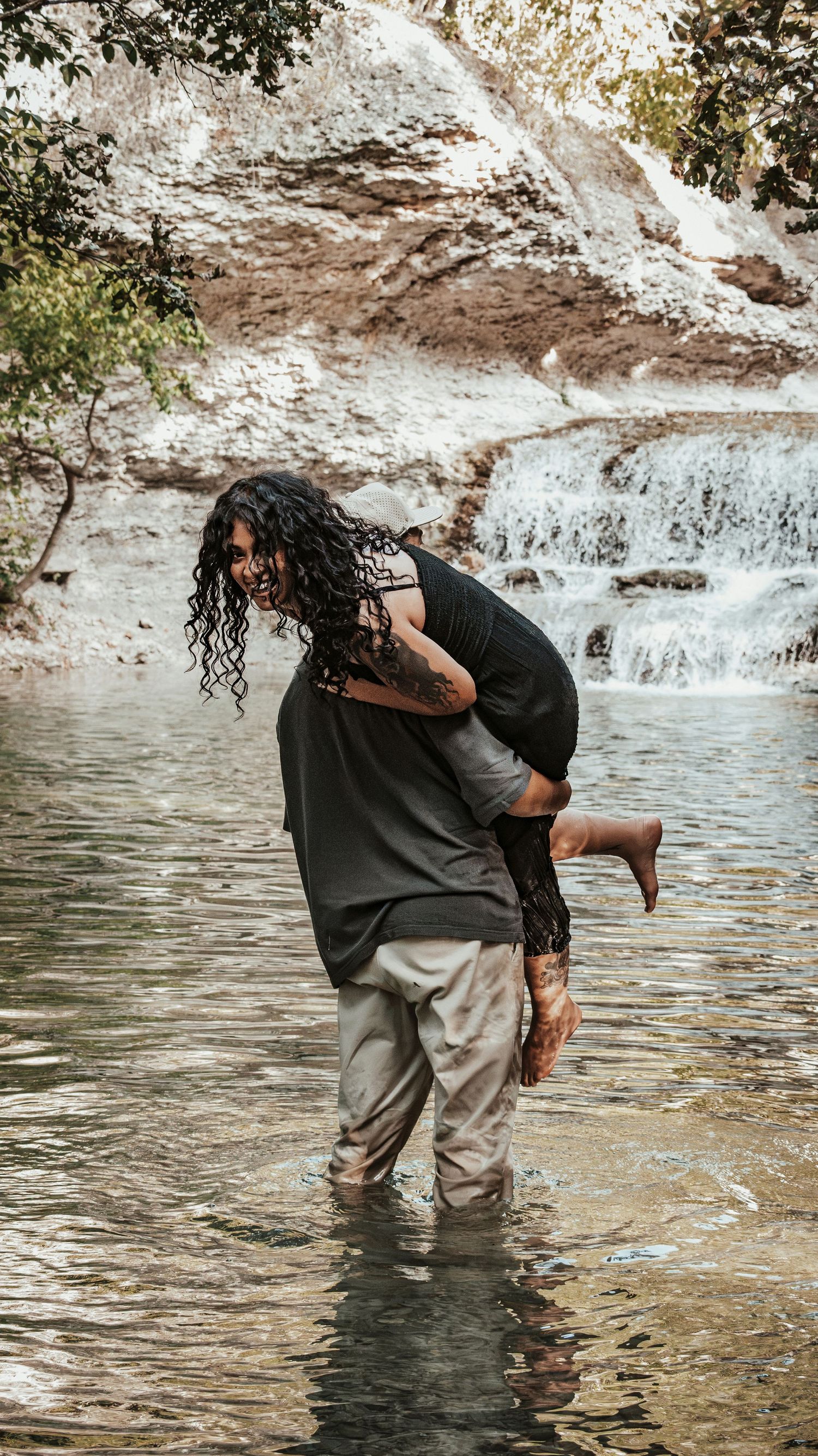 Man carrying woman over his shoulder while wading through water near a cascading waterfall.