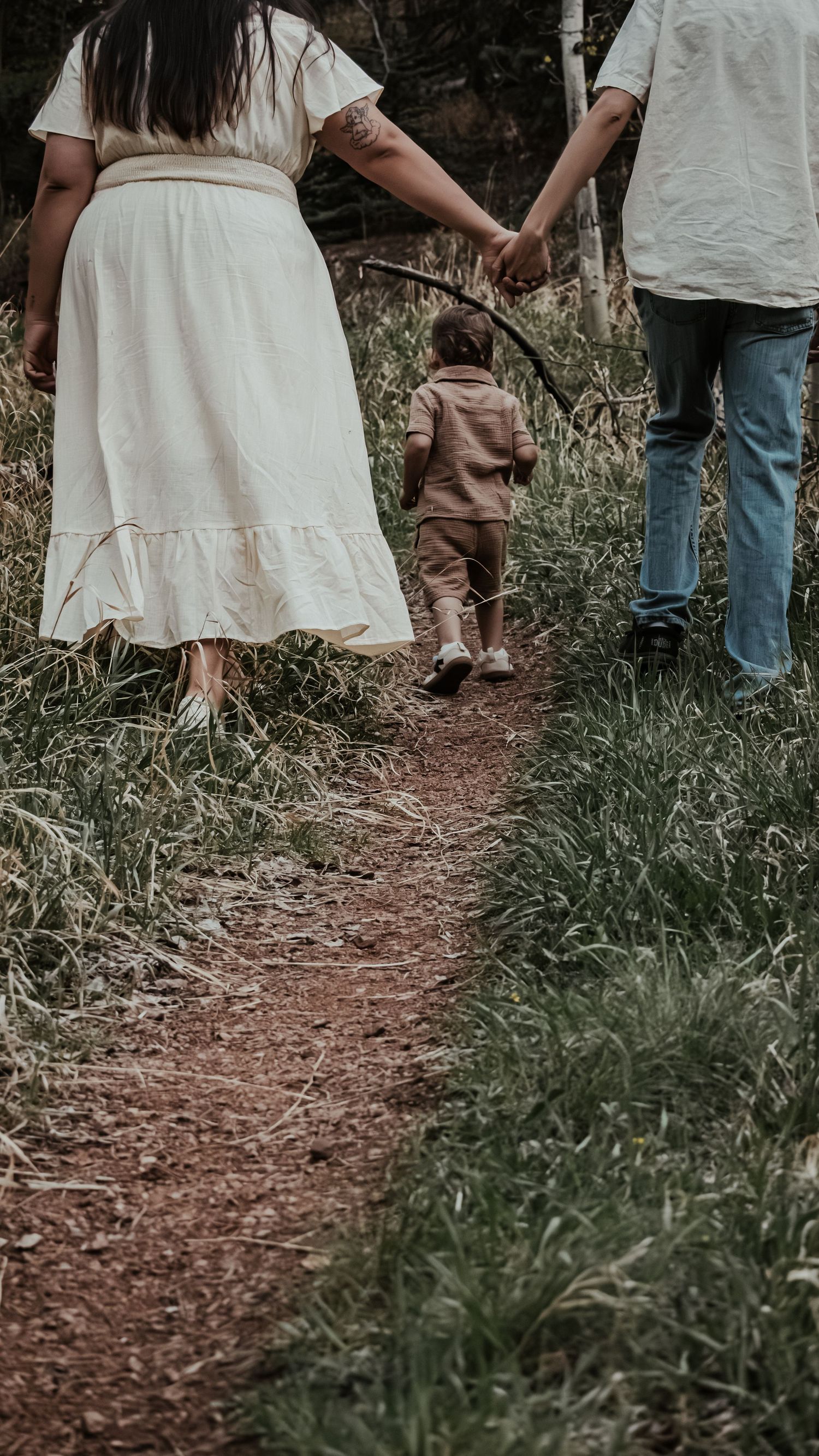 A family of three walks hand-in-hand along a narrow dirt path through green meadow grass.