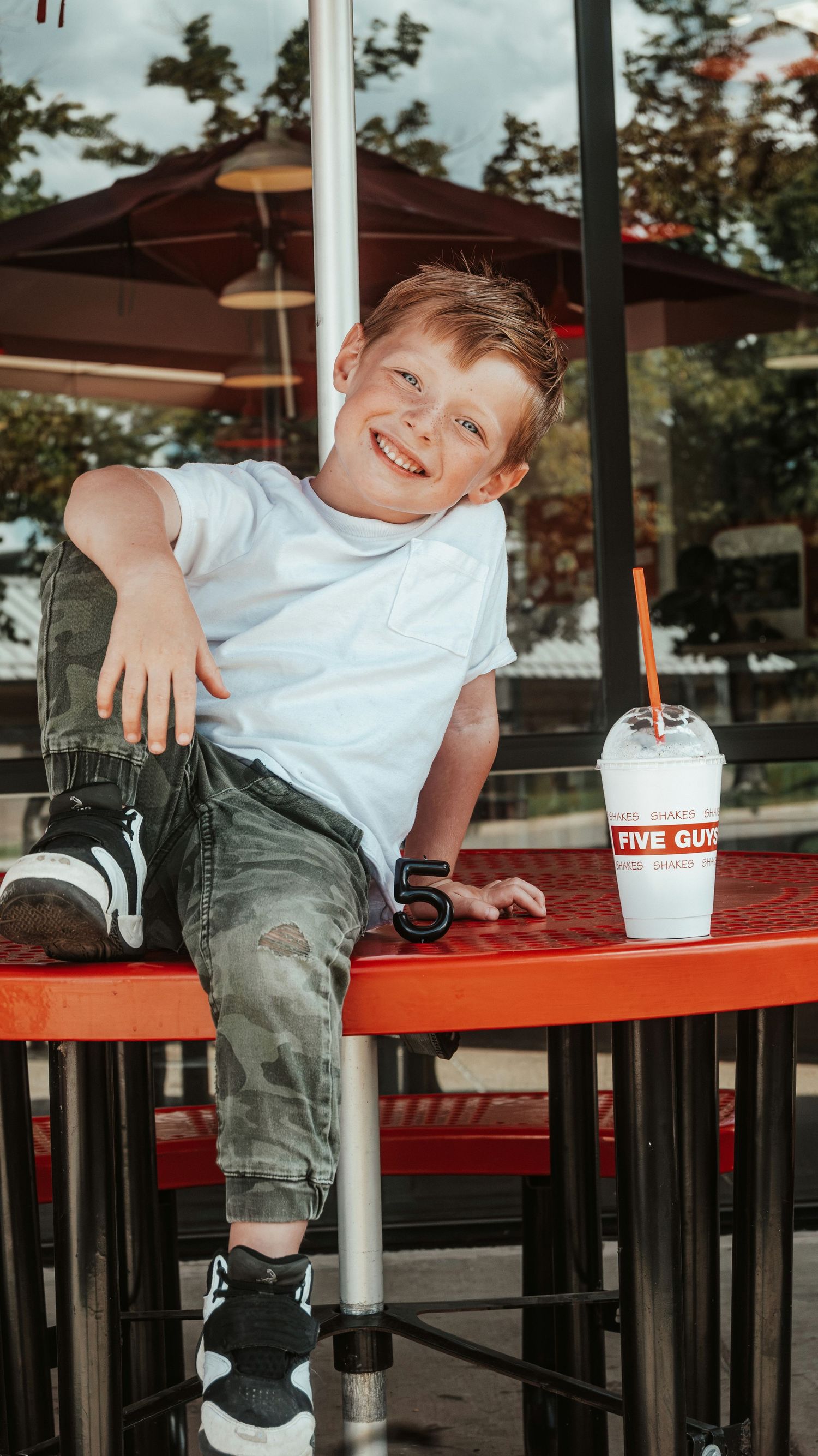 Happy boy sitting on red Five Guys outdoor table smiling with milkshake and number 5.