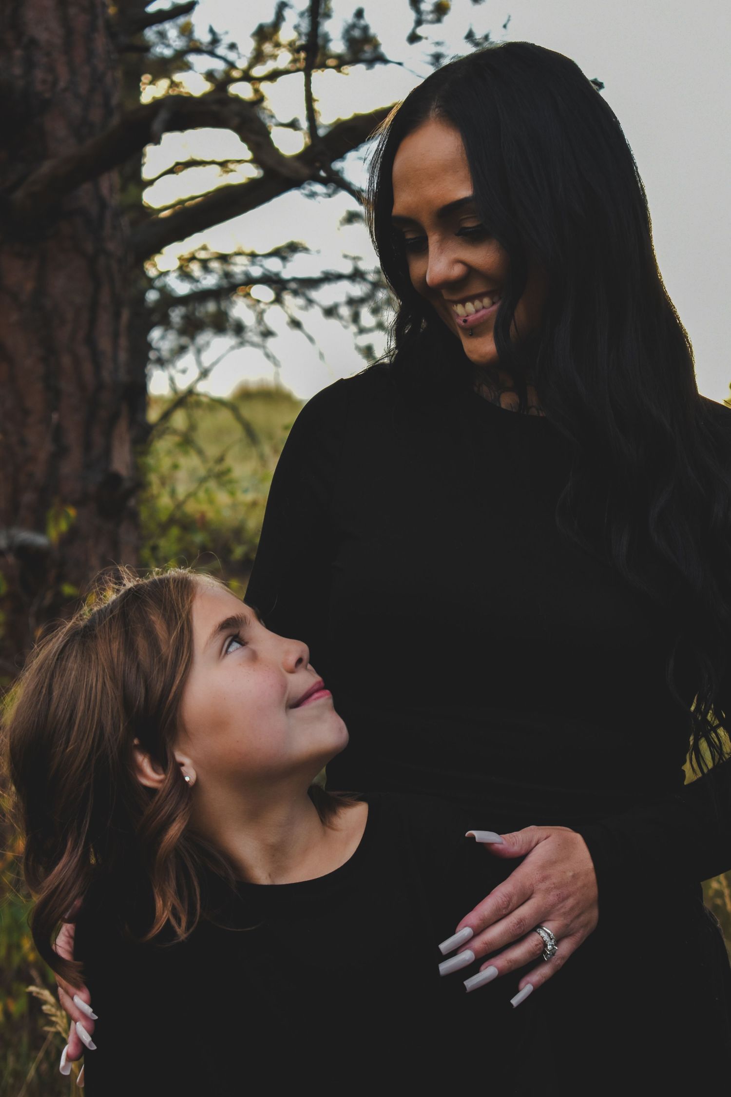 Smiling pregnant woman in black dress with daughter looking up at her near a tree. Family Photographer in Pflugerville, Tx