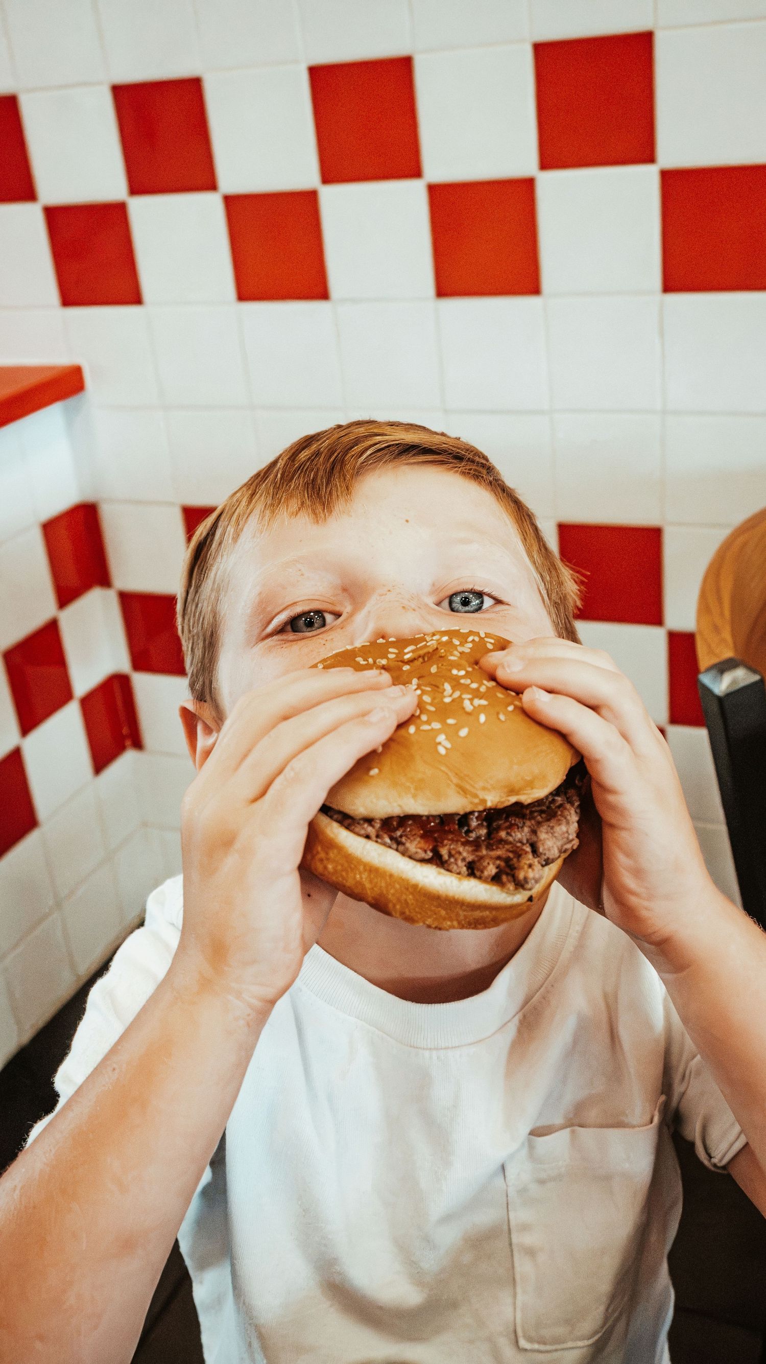 Young boy eagerly biting into a large burger at Five Guys restaurant. Family Photographer in Pflugerville, Tx