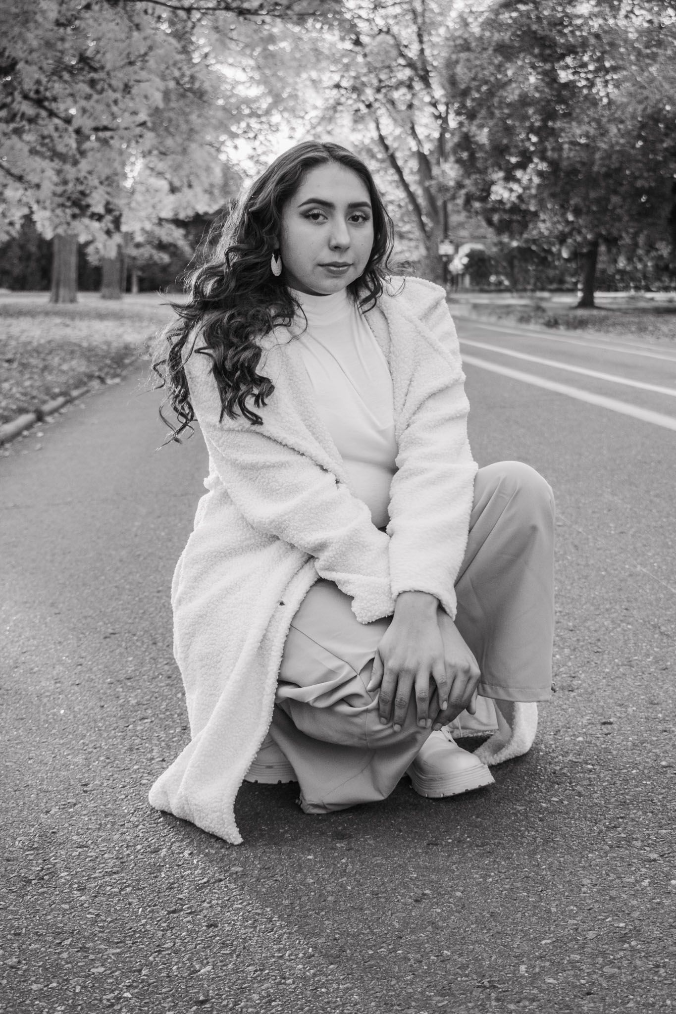 Black and white portrait of young woman in fluffy coat crouching on road in autumn park for senior photos.