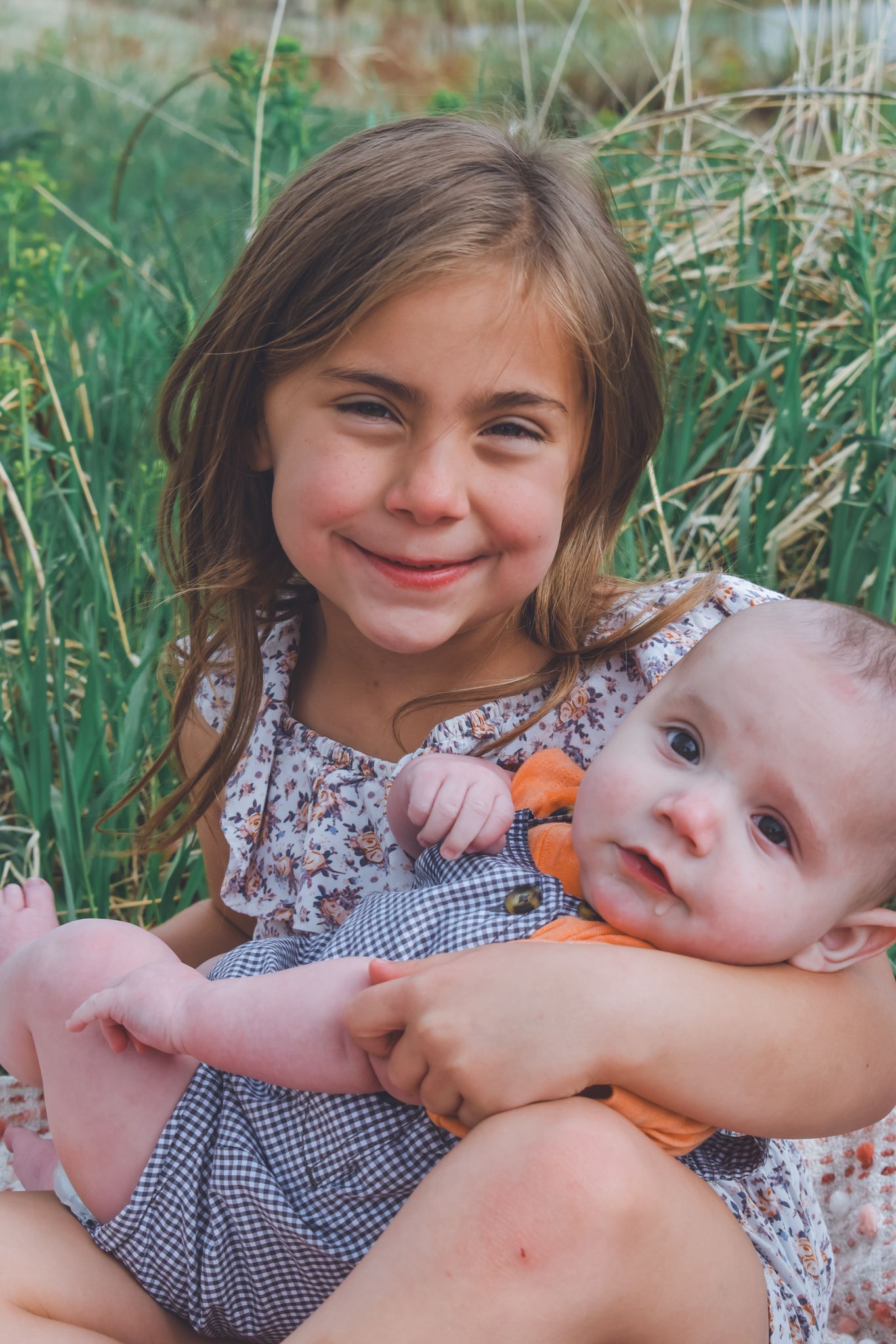 Smiling young girl with long hair gently holding happy baby sibling outdoors in grass.