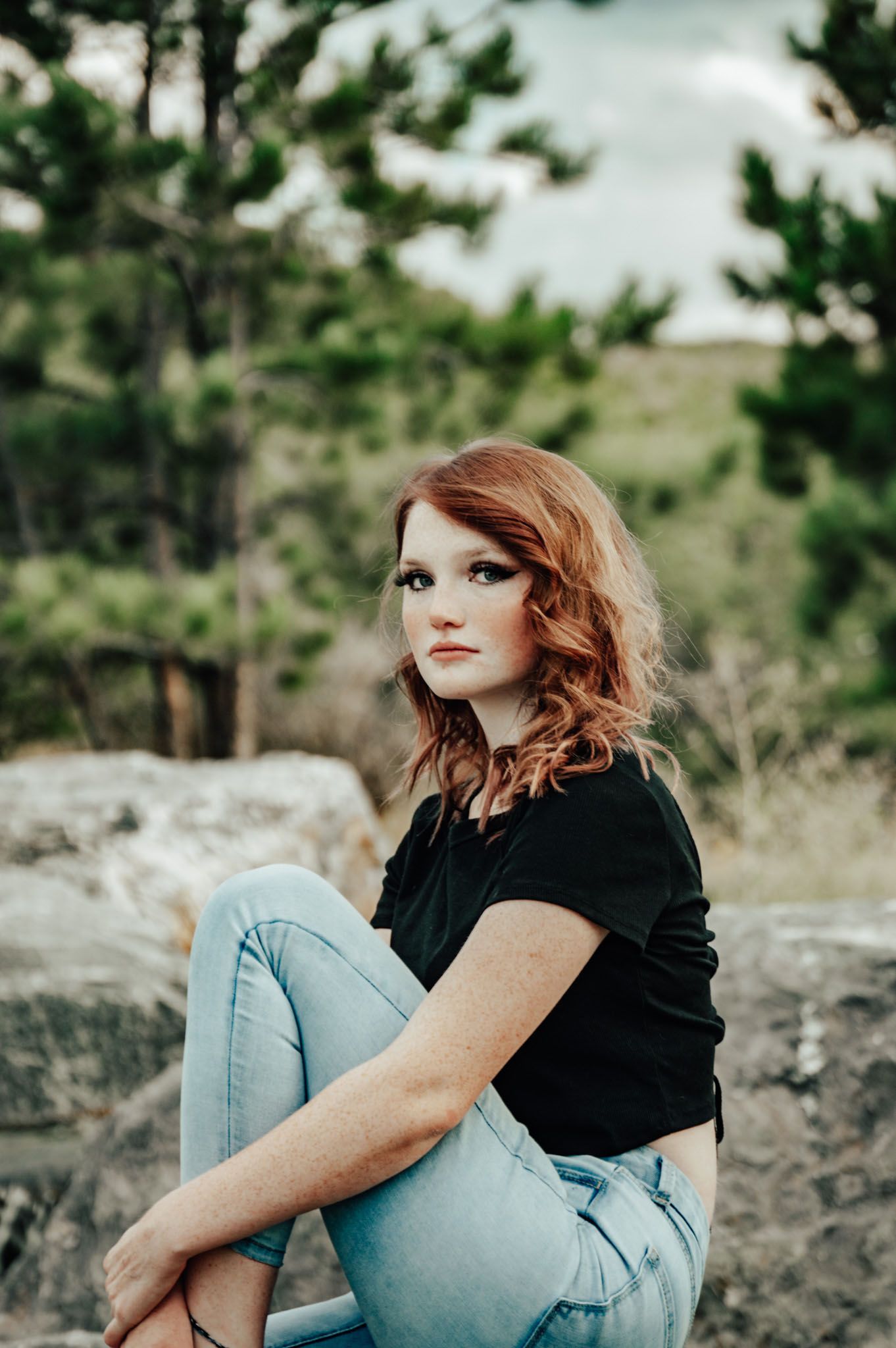 Redhead woman in black tee and light jeans sitting on rocks outdoors with pine trees in background for senior photos.