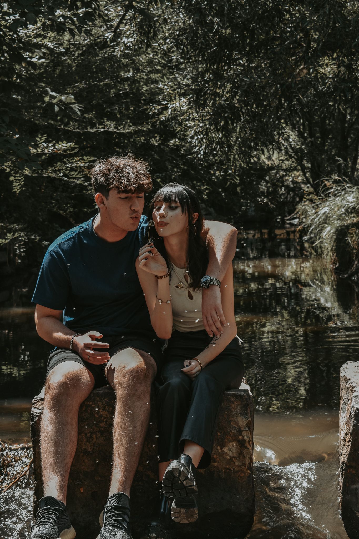 Young couple sits together on a stone ledge near a forest stream, sharing a quiet intimate moment for senior photos.