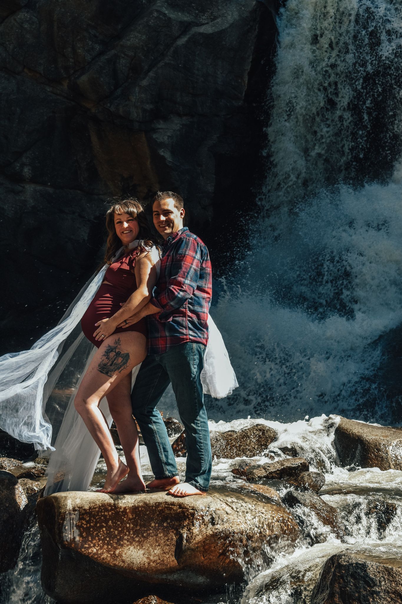 Couple embracing on rocks in front of a waterfall, woman in red dress with veil.  Maternity Photographer in Pflugerville, Tx
