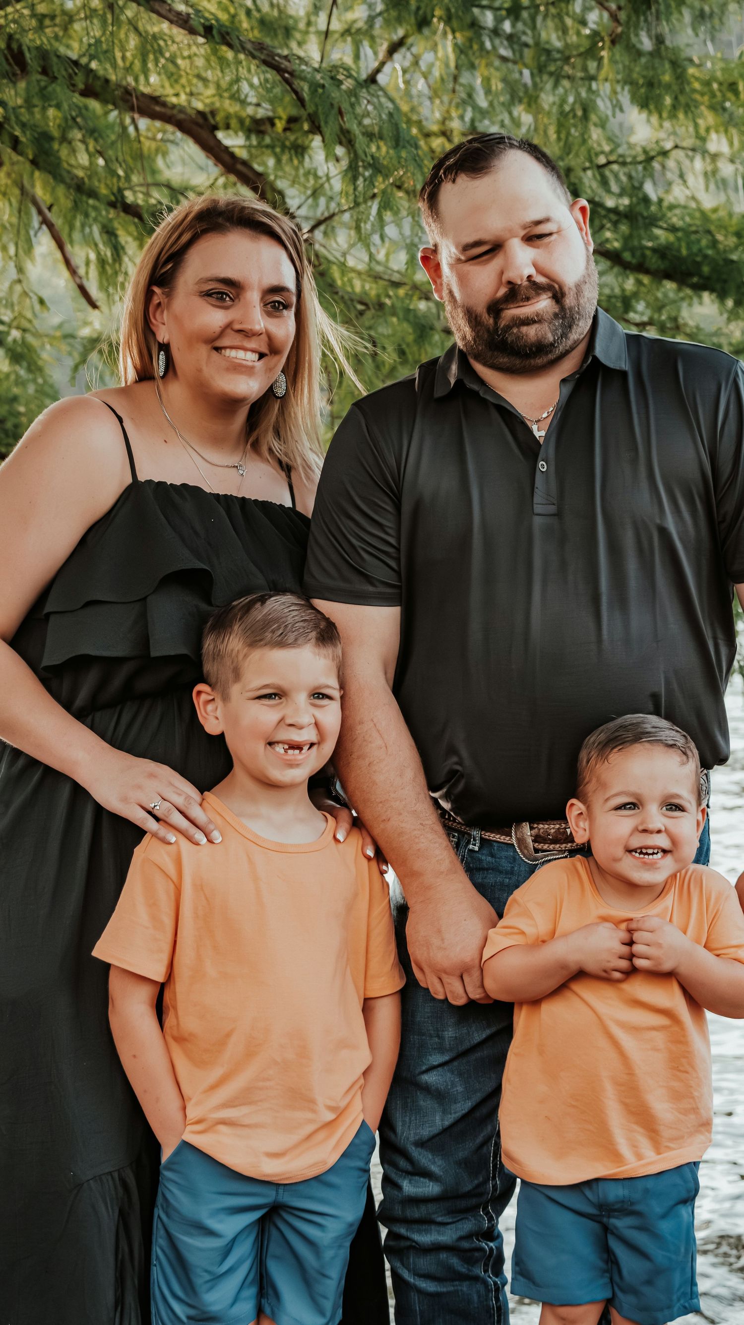 Family of four posing outdoors, parents in black and two young boys in matching orange shirts and blue shorts, smiling.