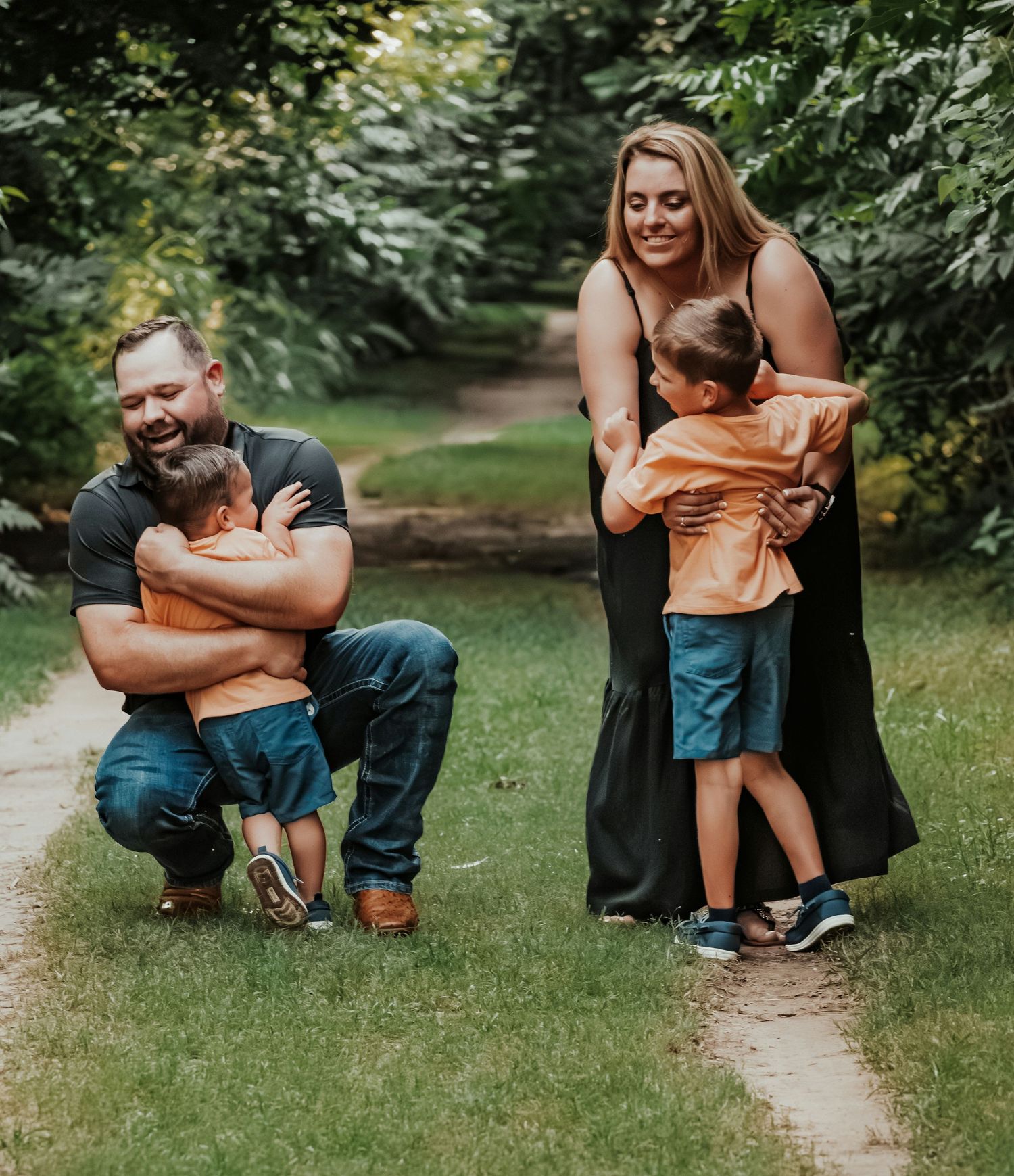 Family of four sharing hugs on a garden path, parents embracing young children outdoors.