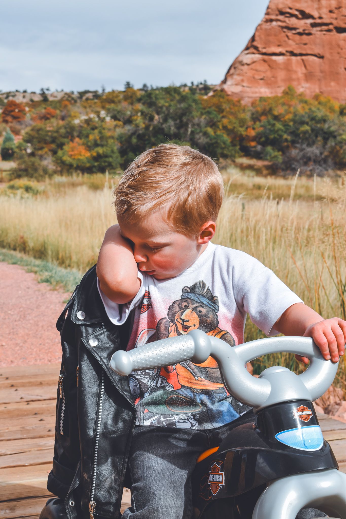 Young boy in graphic tee removing leather jacket while sitting on Harley-Davidson toy motorcycle.