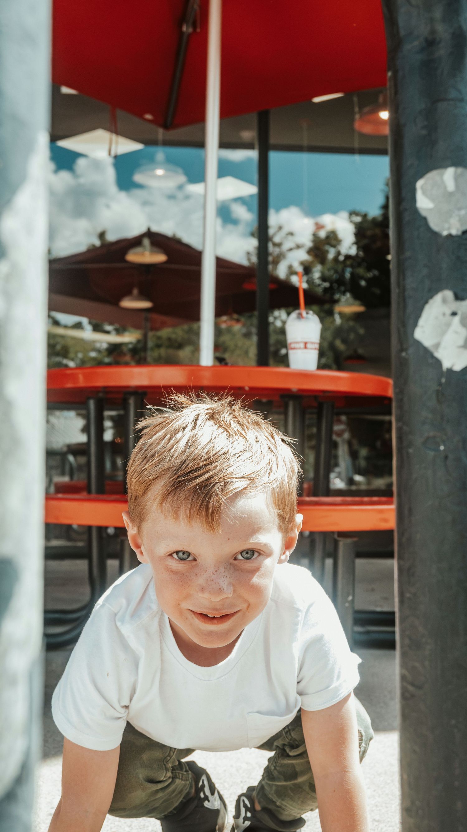 Young boy in white t-shirt crouching playfully outside a Five Guys restaurant on a sunny day.