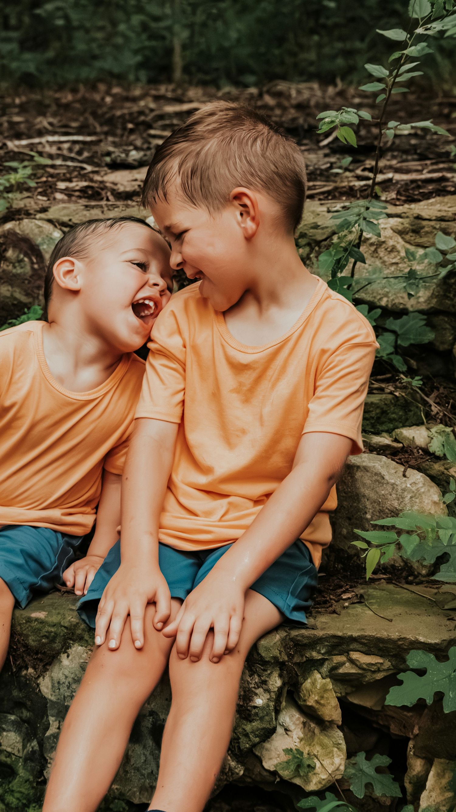 Two young boys in matching orange shirts and teal shorts laugh together while sitting on a stone wall outdoors.