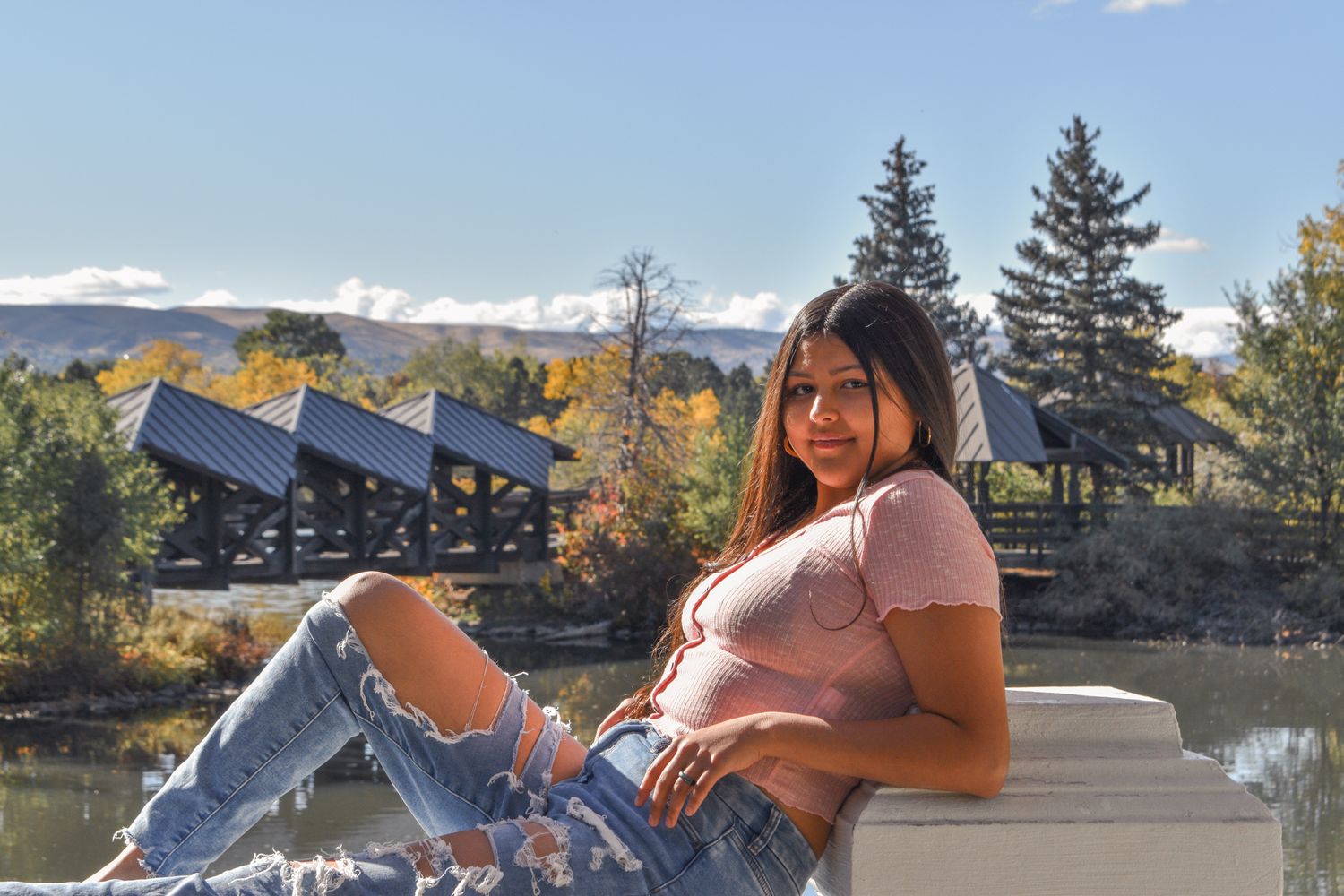 Young woman in pink top and ripped jeans sits on ledge with mountain lake view for senior photos.