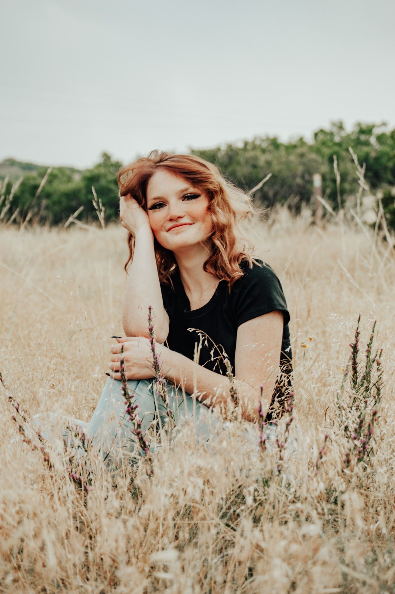 Smiling redhead woman with freckles sitting in golden dry grass field wearing black tee and light jeans for senior photos.