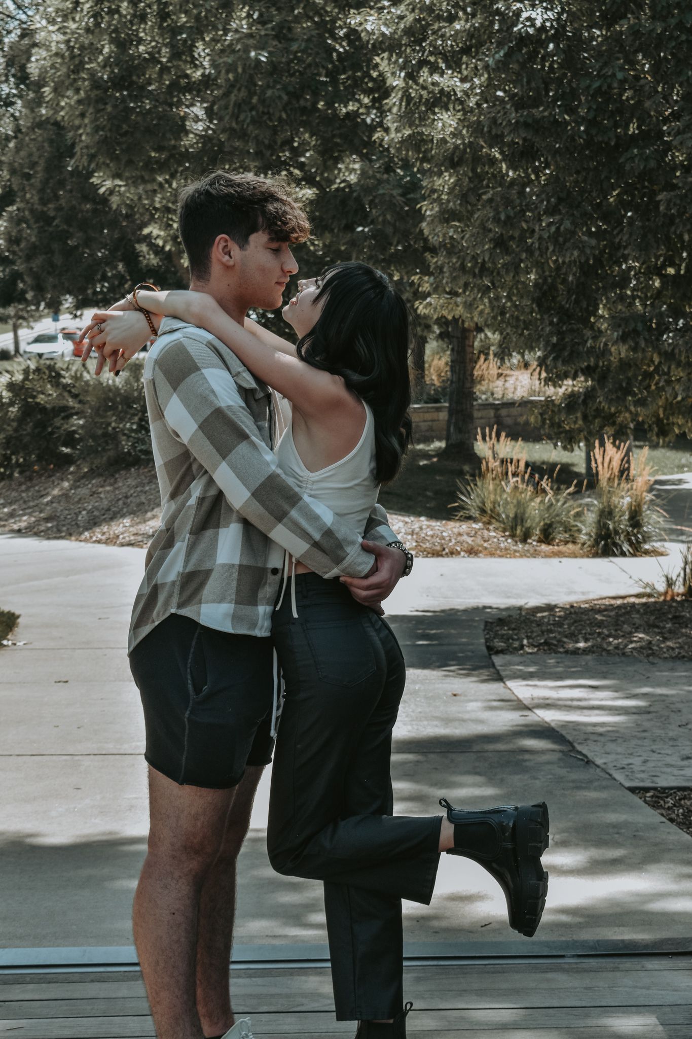 Young couple shares a romantic kiss while embracing outdoors on a sunny tree-lined pathway for senior photos.