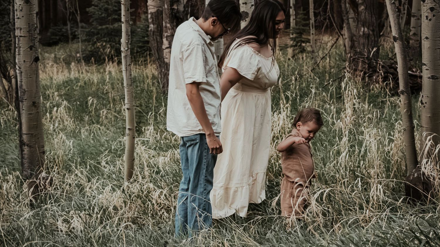 Two parents stand with their young toddler child among tall grass and birch trees in nature.