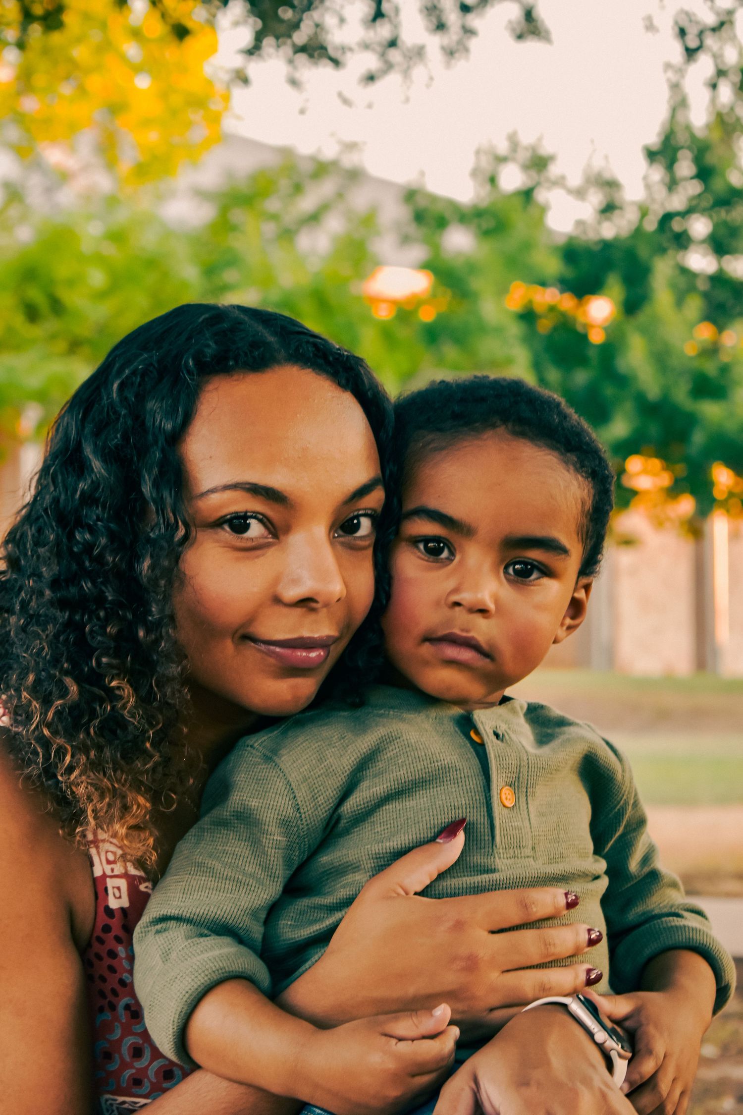 A loving African American mother embraces her young son outdoors in a park during warm golden hour light.
