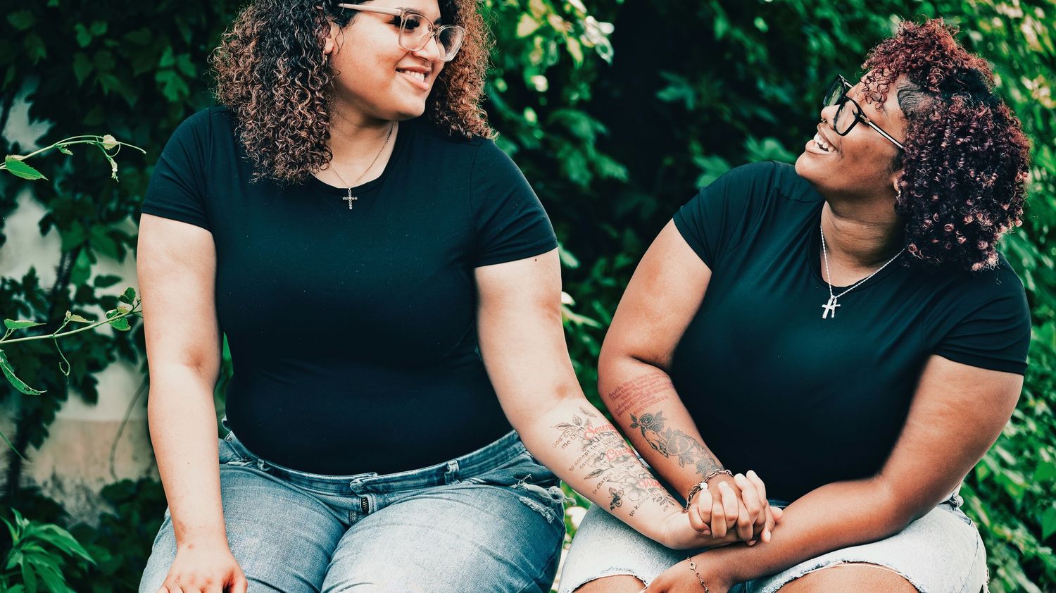 Two smiling women in black tops holding hands while sitting outdoors among green foliage for senior photos.