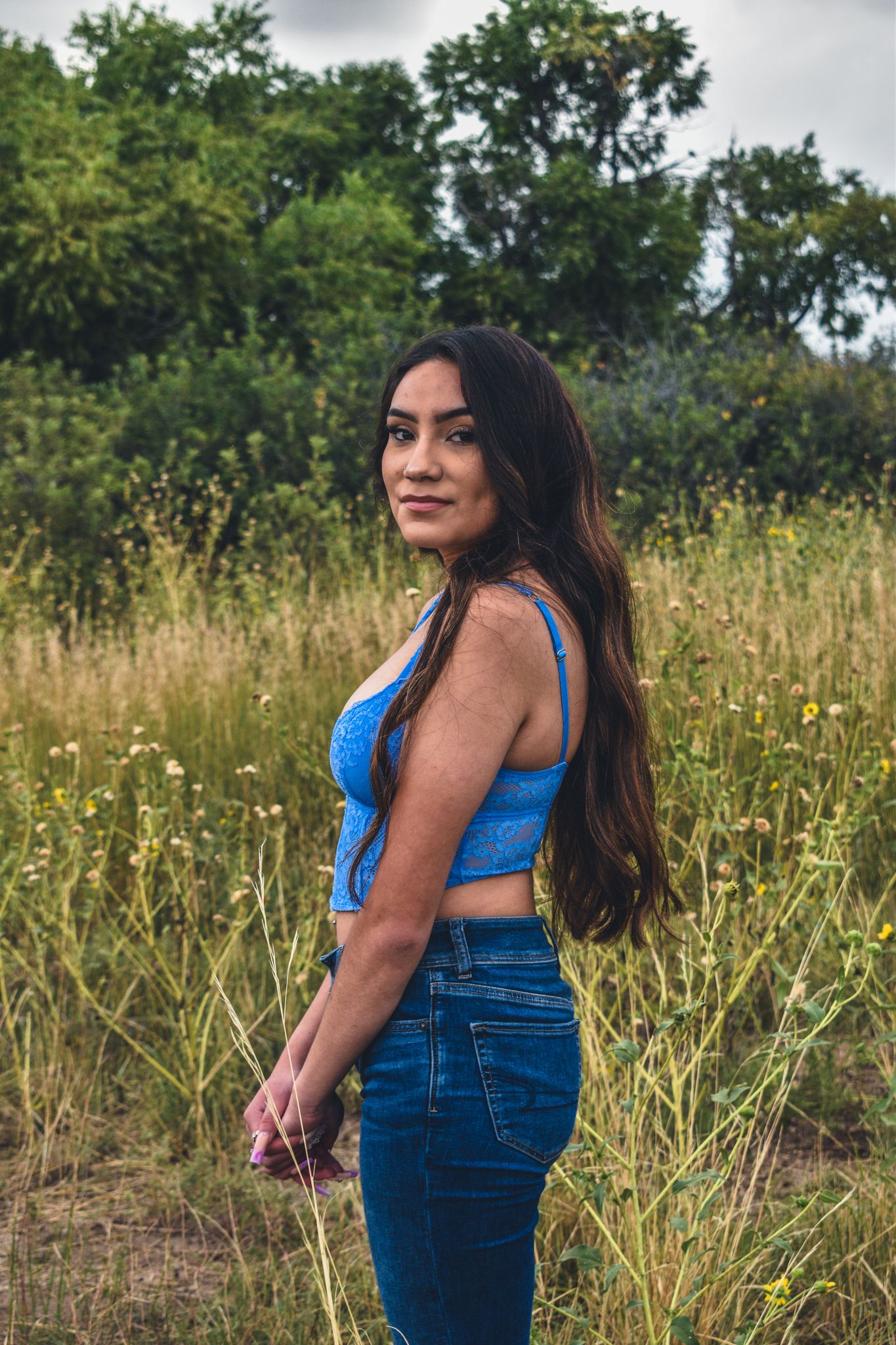 Young woman in blue crop top and jeans standing in a wildflower field, smiling over shoulder for senior photos.
