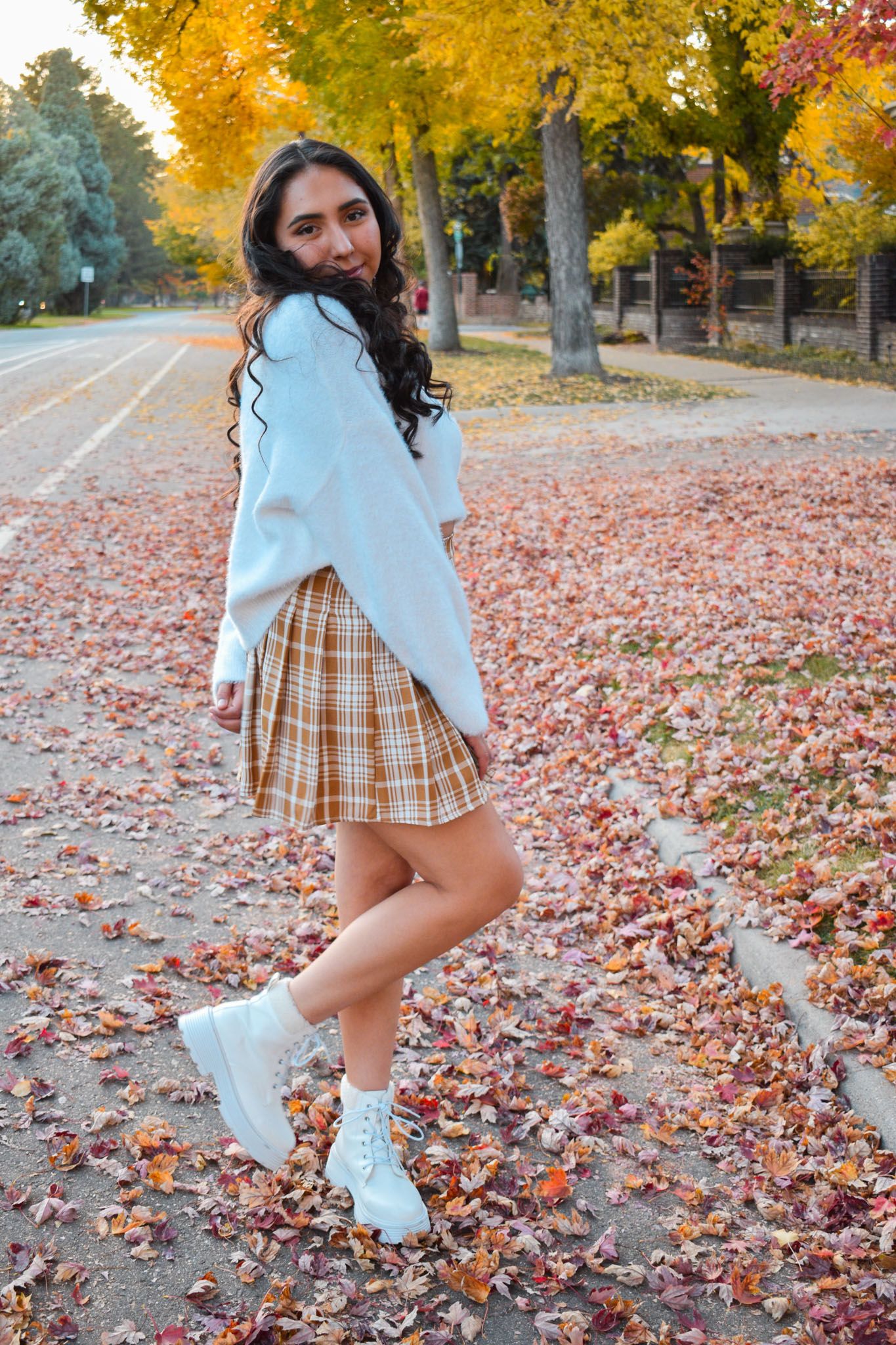 Young woman in white sweater and plaid skirt posing among fallen autumn leaves on sidewalk for senior photos.