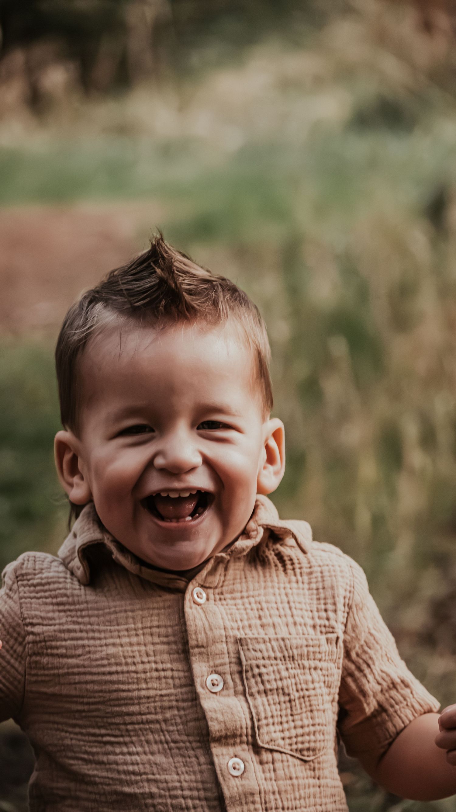 Happy laughing toddler boy with a mohawk haircut wearing a beige linen shirt in a natural outdoor setting.