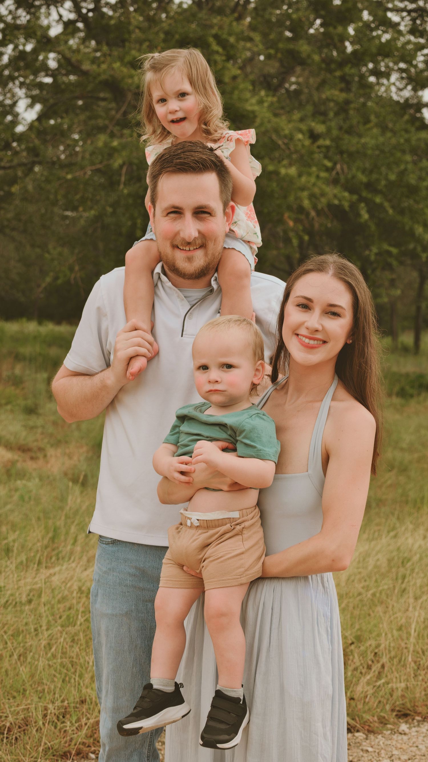 Cheerful family of four posing in a green field with toddler on dad's shoulders.