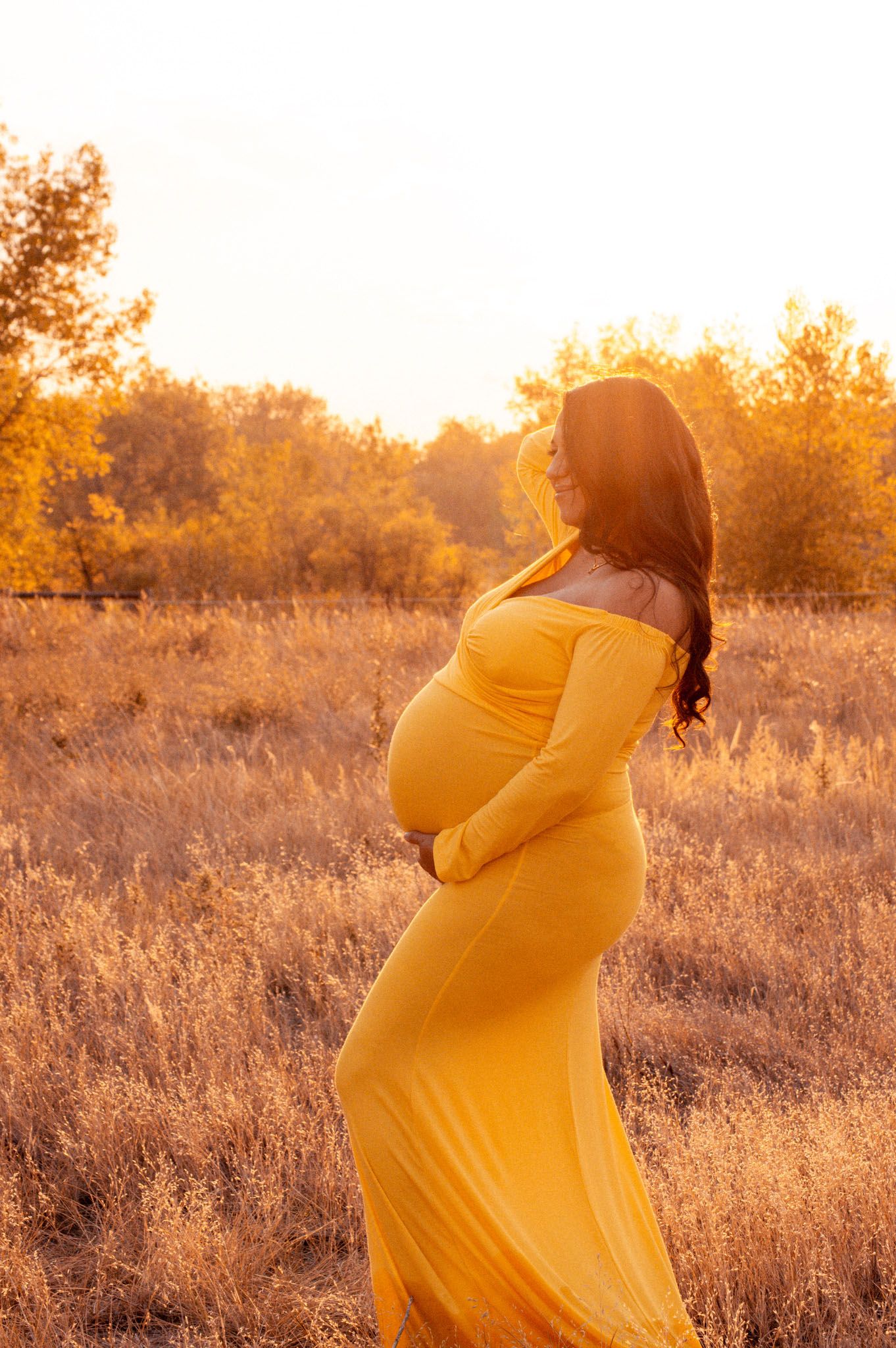 Glowing pregnant woman in yellow maxi dress standing in golden autumn field.